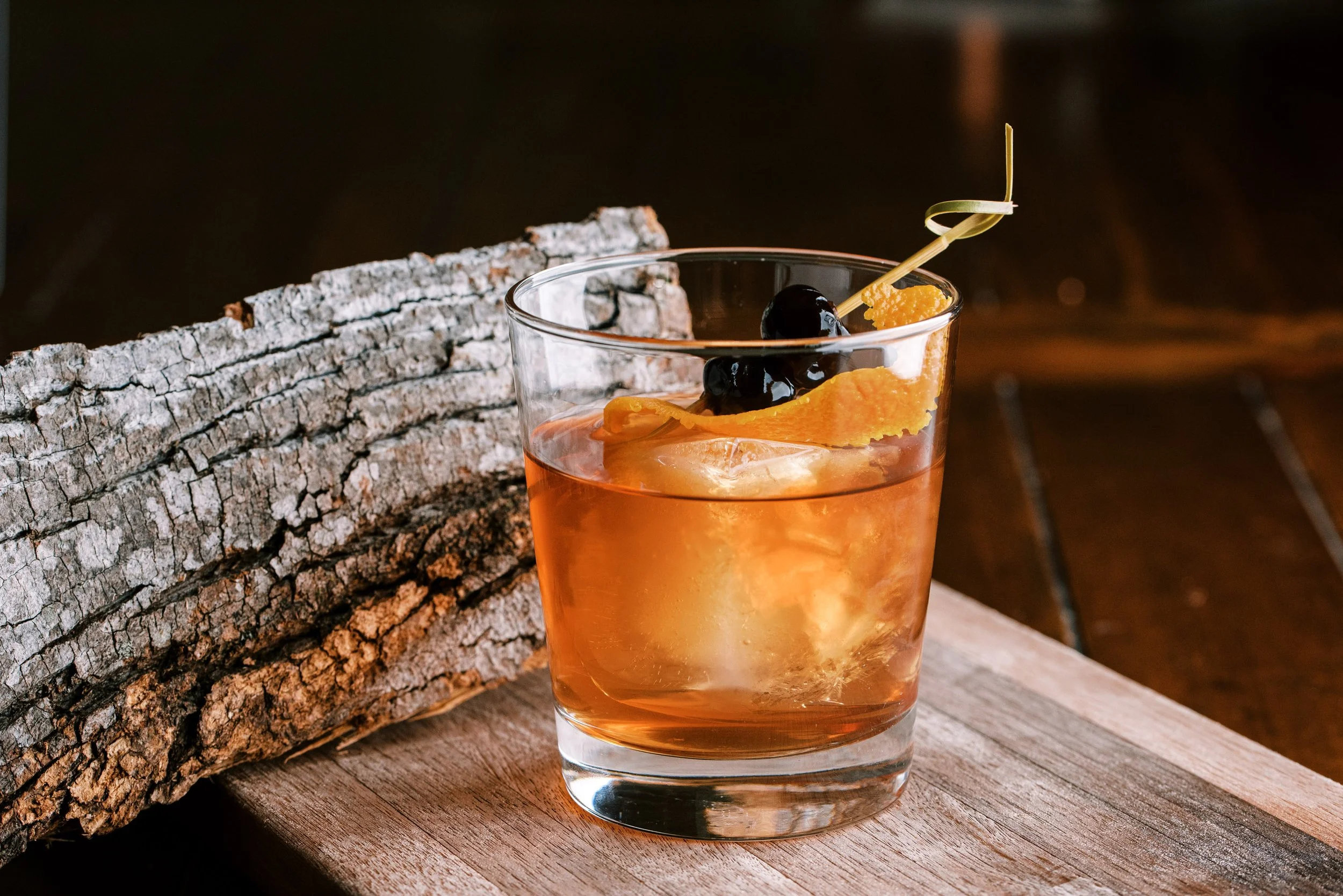 Amber whiskey cocktail with orange peel and cherry garnish on a rustic wooden surface, drink photography by Cody Myers