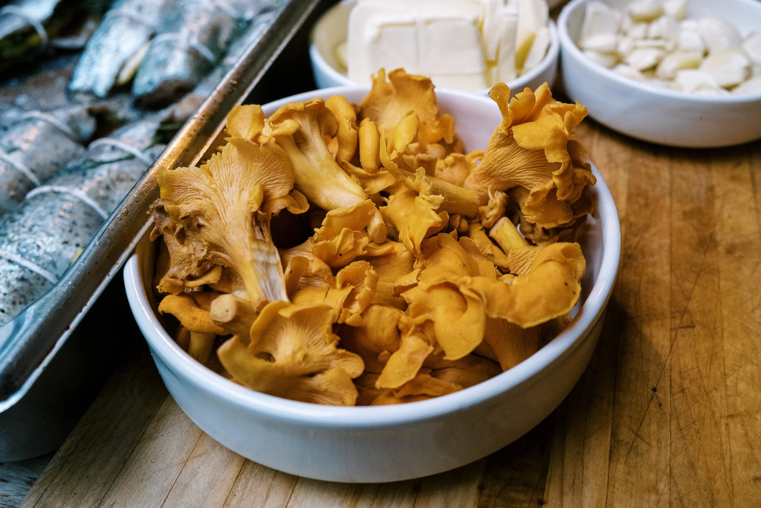 Fresh chanterelle mushrooms in a white ceramic bowl on a wooden prep surface, foraging food photography by Cody Myers
