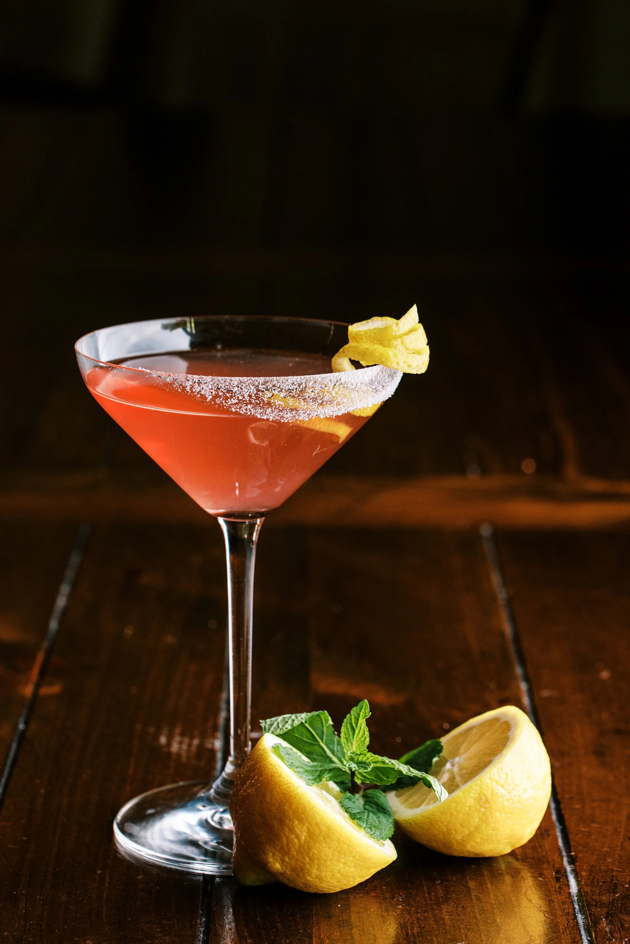 Pink citrus cocktail in a coupe glass with lemon garnish on a dark wood bar, drink photography by Cody Myers
