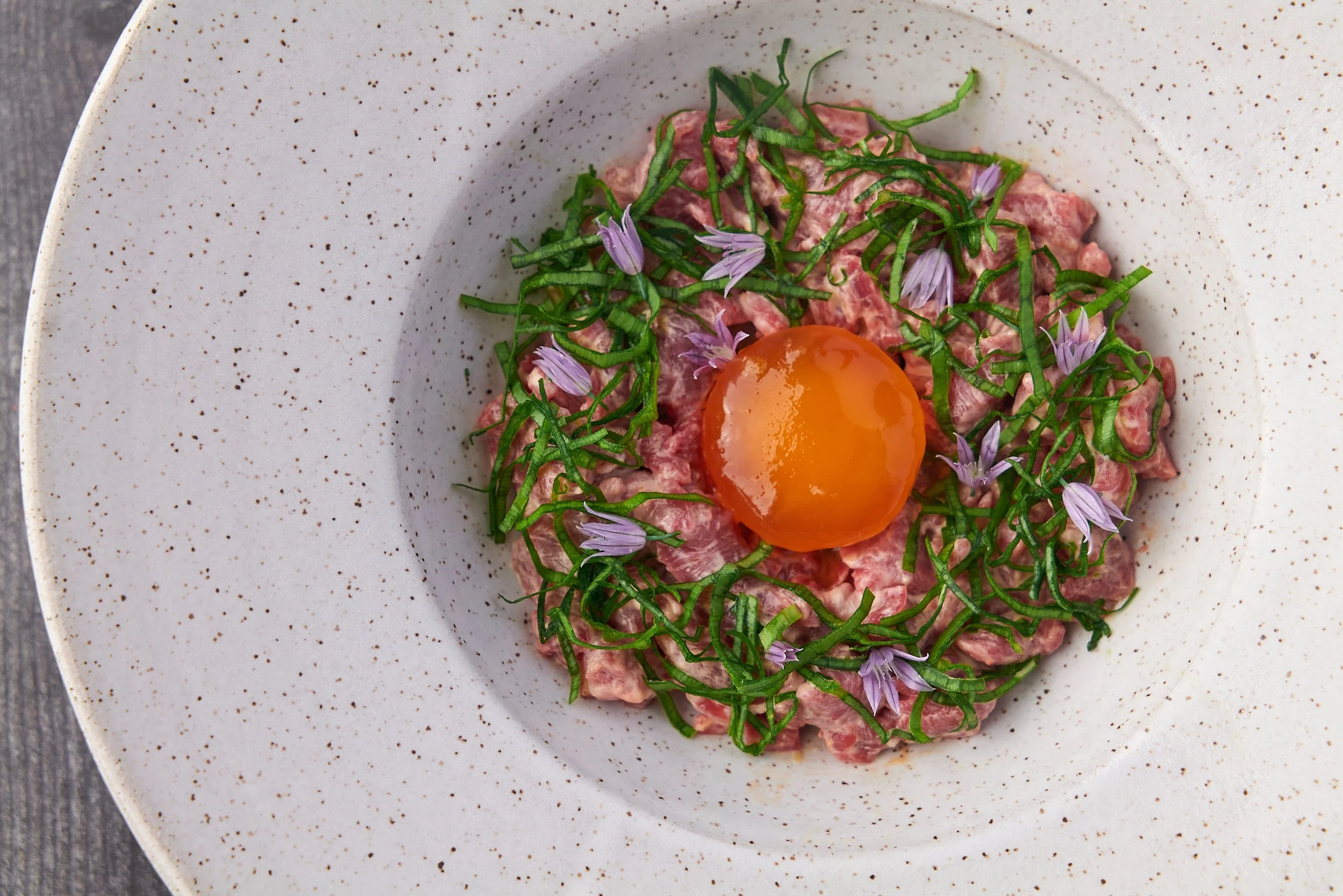 Steak tartare with raw egg yolk, fresh herbs and edible flowers on a white ceramic plate, fine dining food photography by Cody Myers