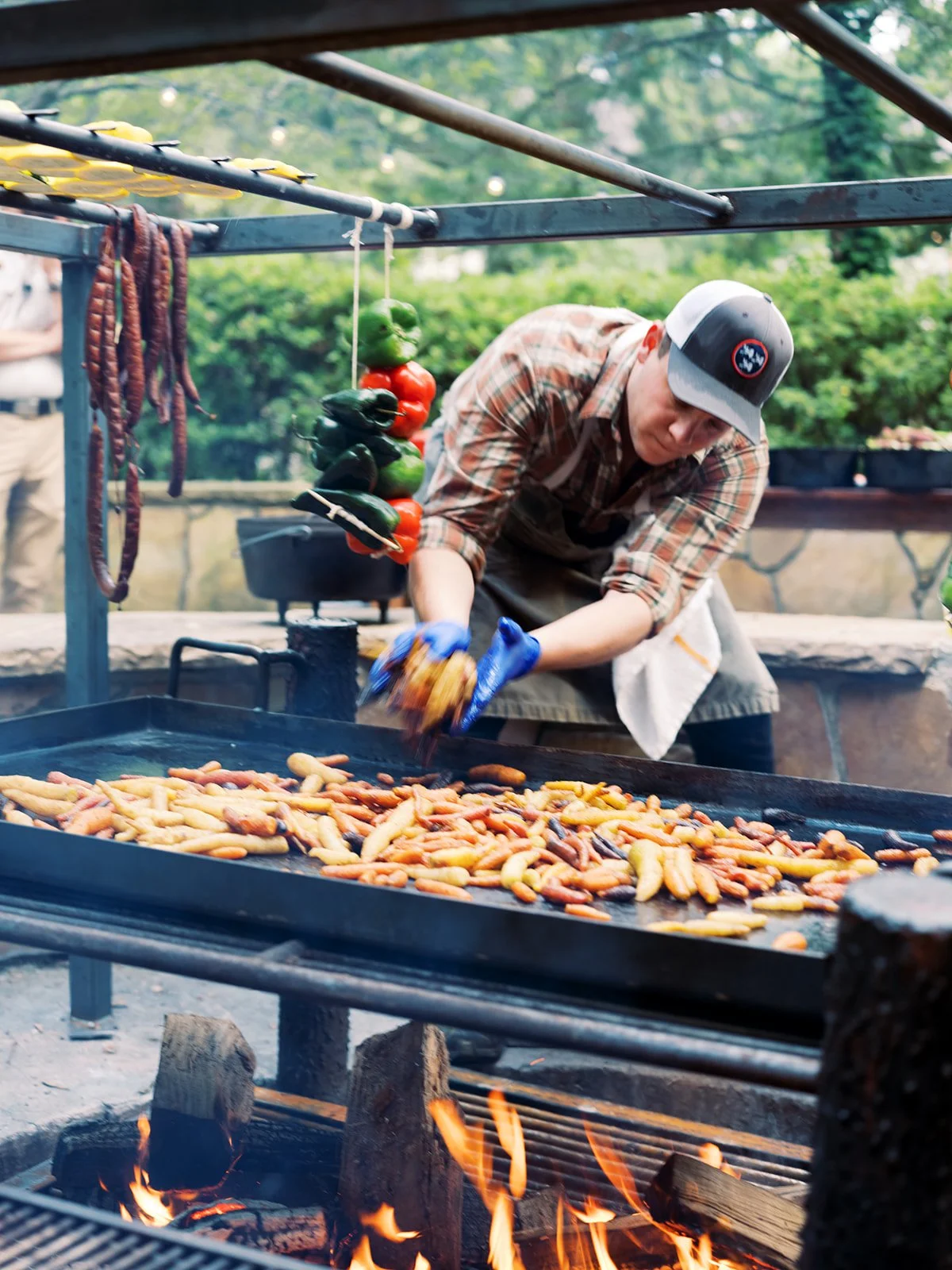 Chef tending an open-flame grill loaded with food at an outdoor cooking event, lifestyle food photography by Cody Myers