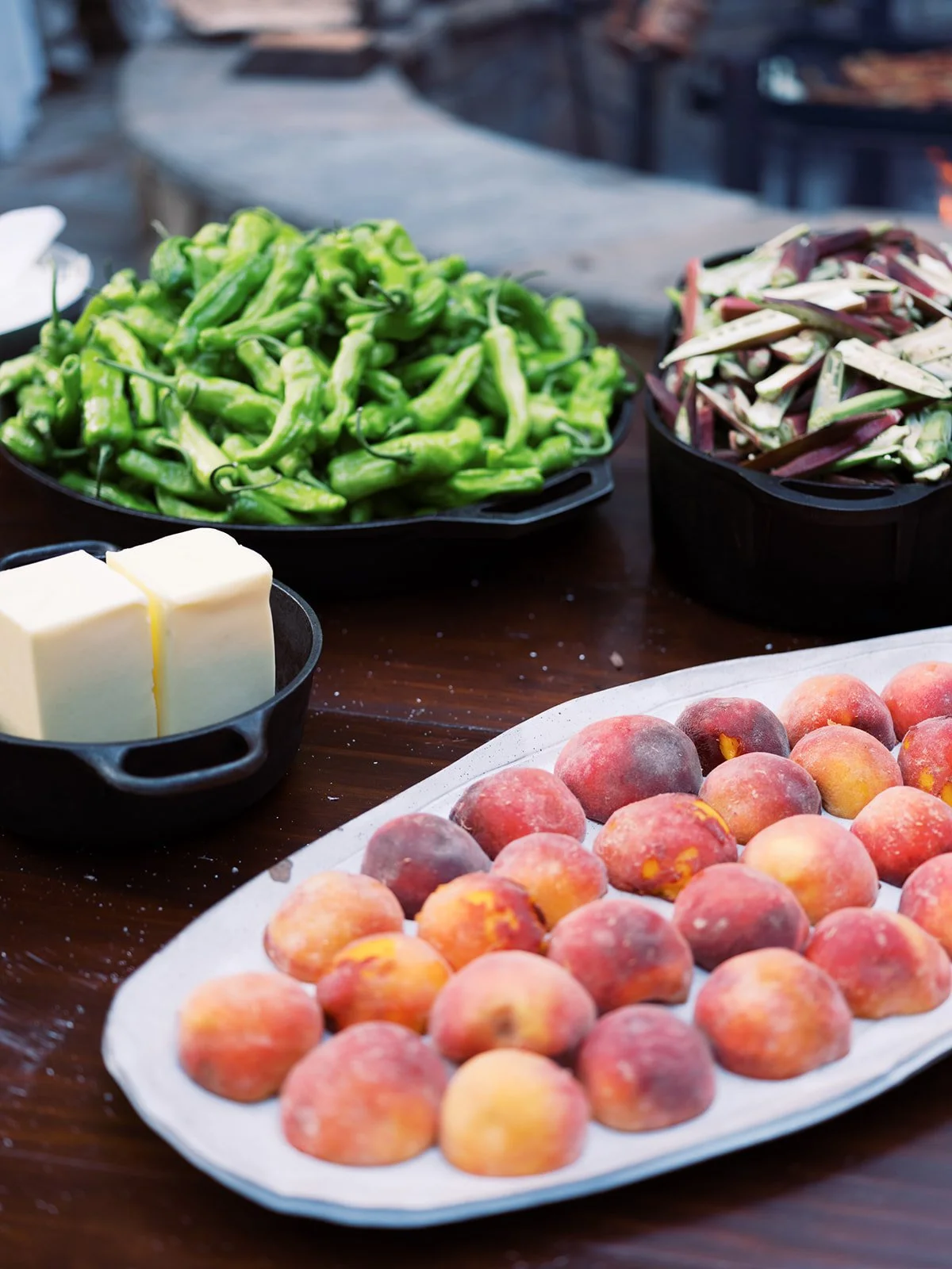 Fresh peaches on a serving platter alongside shishito peppers and grilled vegetables at an outdoor dinner, food photography by Cody Myers