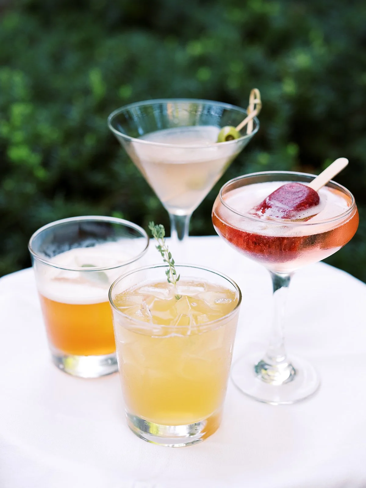 Three handcrafted cocktails displayed on a table at an outdoor garden event, drink photography by Cody Myers