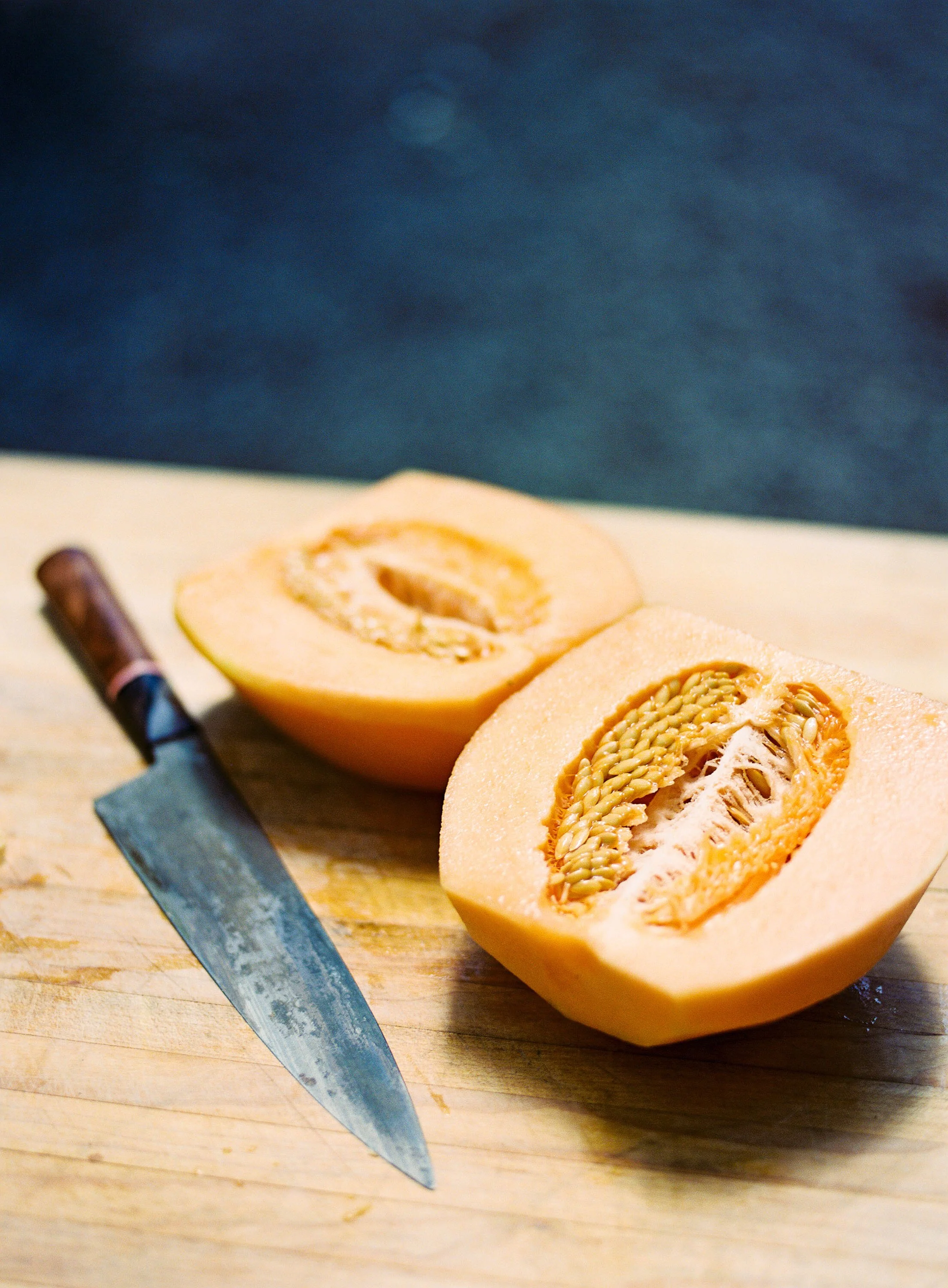 Halved cantaloupe melon with a chef's knife on a wooden cutting board, food photography by Cody Myers