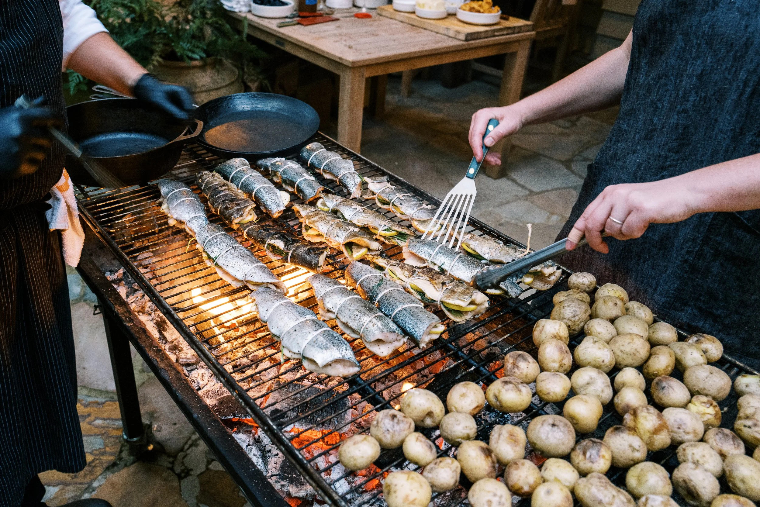 Whole fish and potatoes grilling over open flames at an outdoor dinner event, food photography by Cody Myers