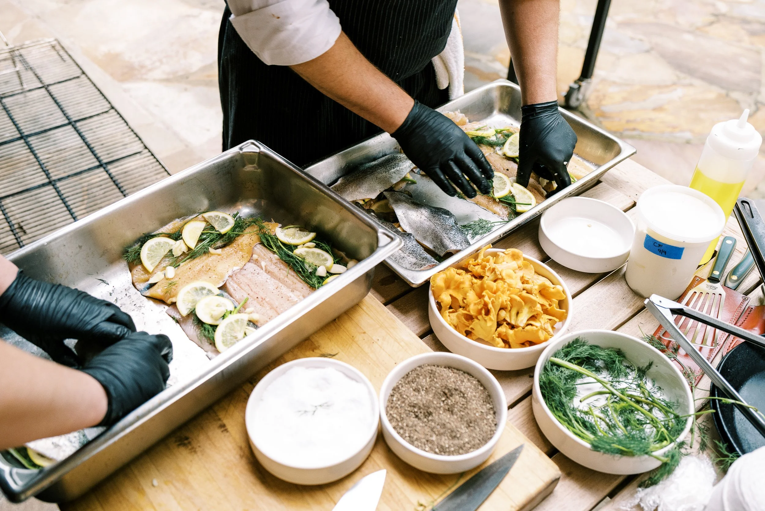 Chefs in black gloves prepping fish and sides in sheet pans at an outdoor cooking station, food photography by Cody Myers