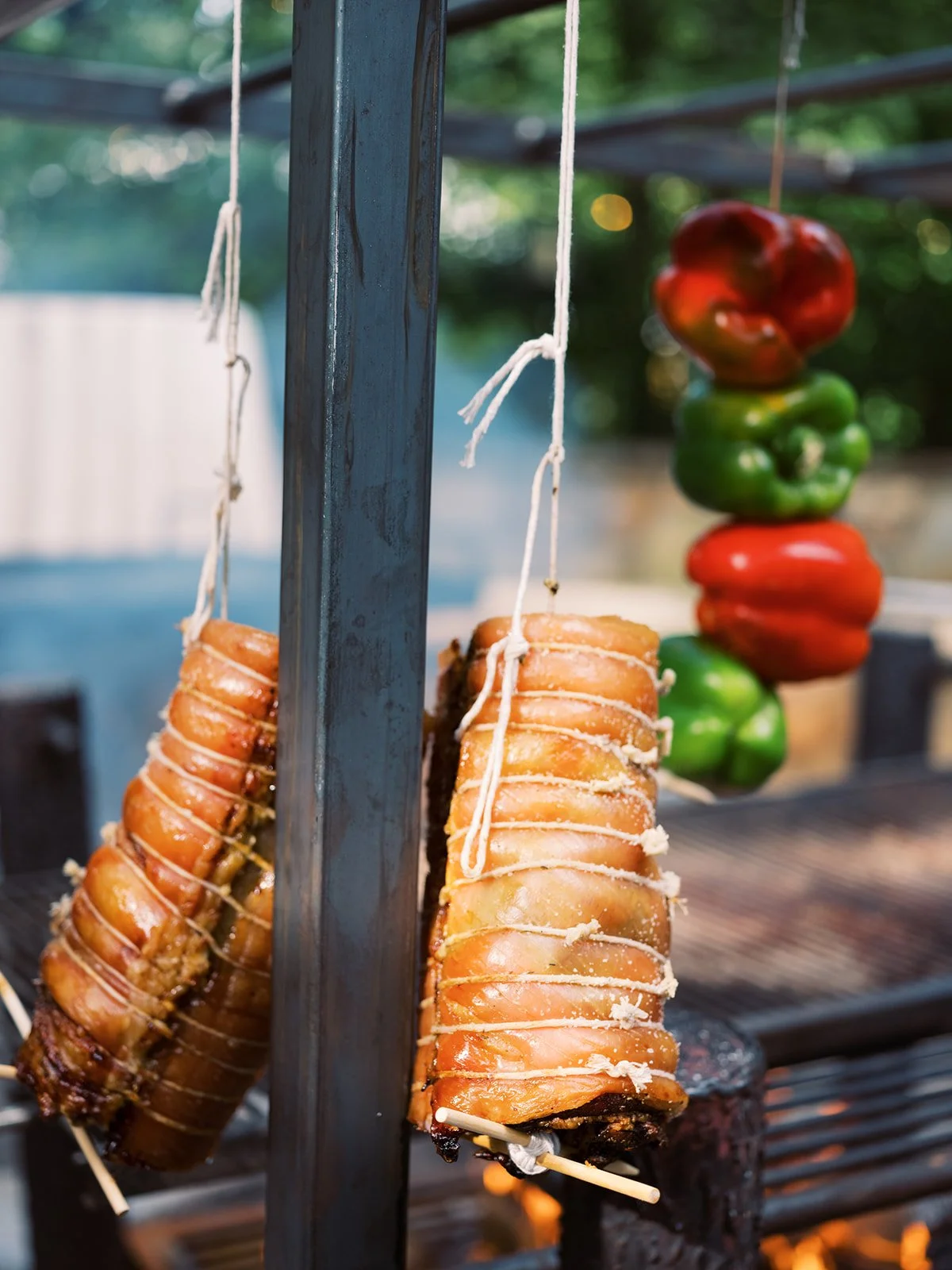 Whole chicken trussed and hanging on an outdoor rotisserie grill with colorful peppers, food photography by Cody Myers