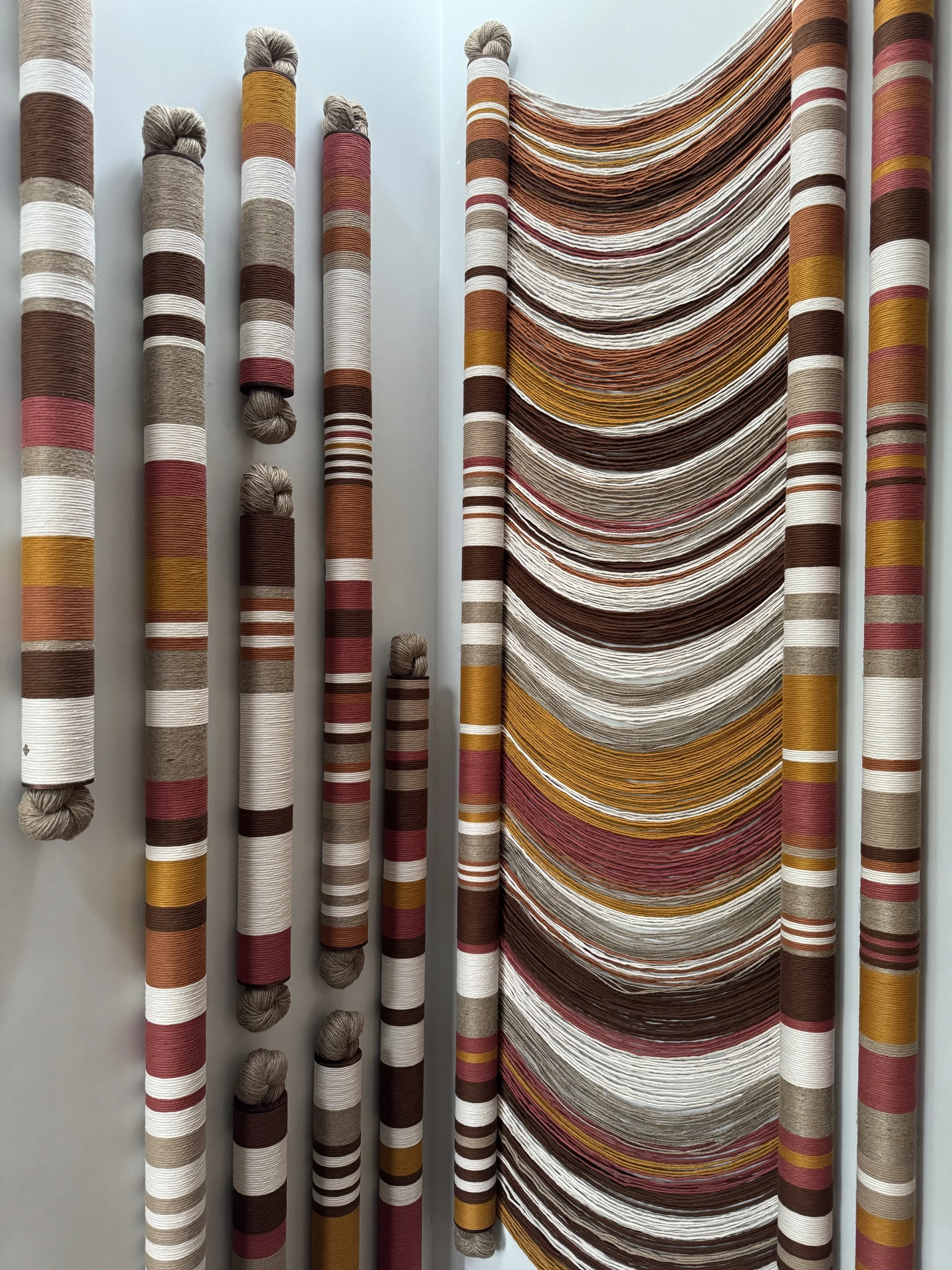 Varias bobinas de hilo en tonos de marrón, beige, blanco, con algunos en rojo, amarillo y naranja, y una cortina hecha con hilos en tonos similares, con detalles en ramas de colores y textura de tela.