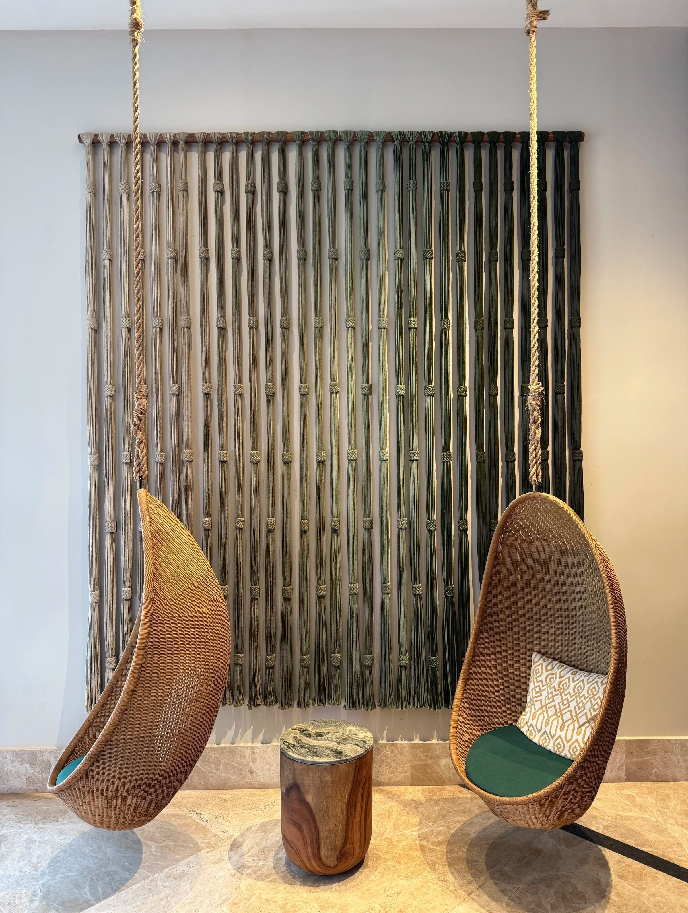 Dos sillas de mimbre con respaldo alto y asiento cubierto con cojín verde, frente a una cortina decorativa de cuerdas en tonos beige, gris y negro, en una pared blanca.