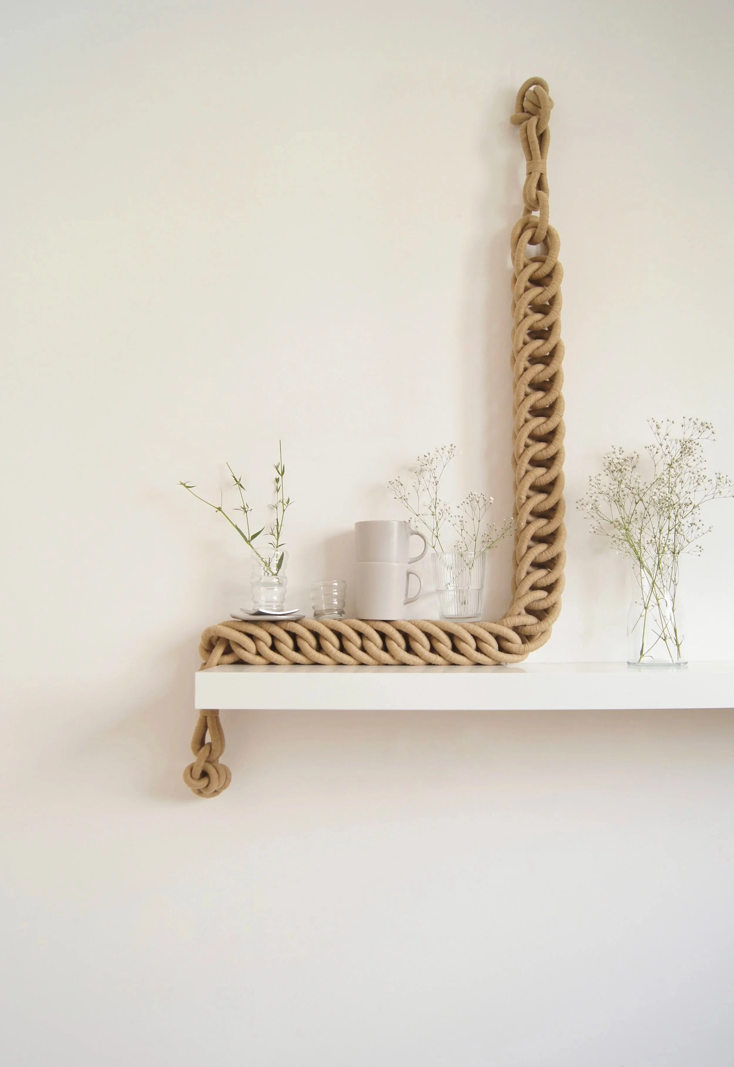 Espejo decorativo de marco grueso de cuerda trenzada, con estantes blancos que sostienen tazas blancas y vasos, decorados con pequeñas ramas y flores secas.