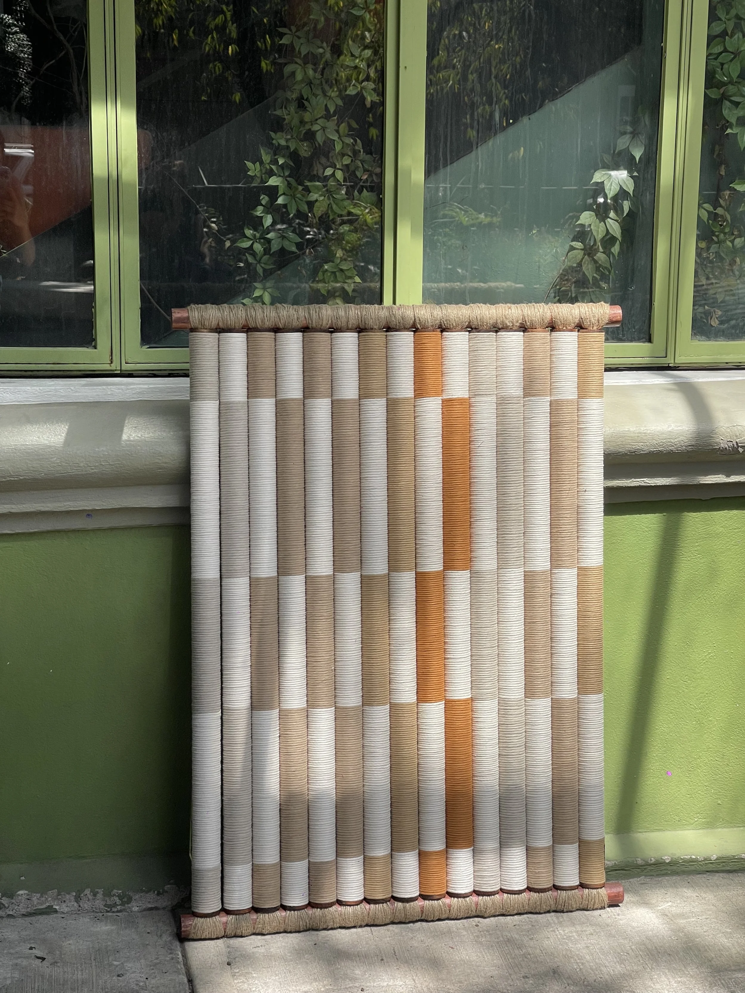 Pantalla decorativa de tubos de bambú en tonos beige, naranja y marrón, colocada frente a una ventana de marco verde.