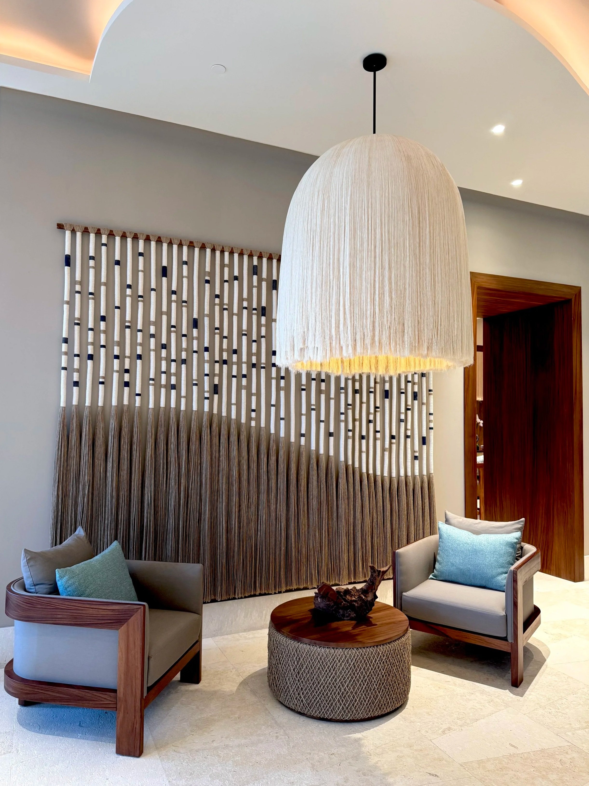 Sala con sillones de tela gris y cojines, una mesa redonda de madera con escultura de madera, cortinas decorativas en la pared y lámpara colgante grande de fibra natural.