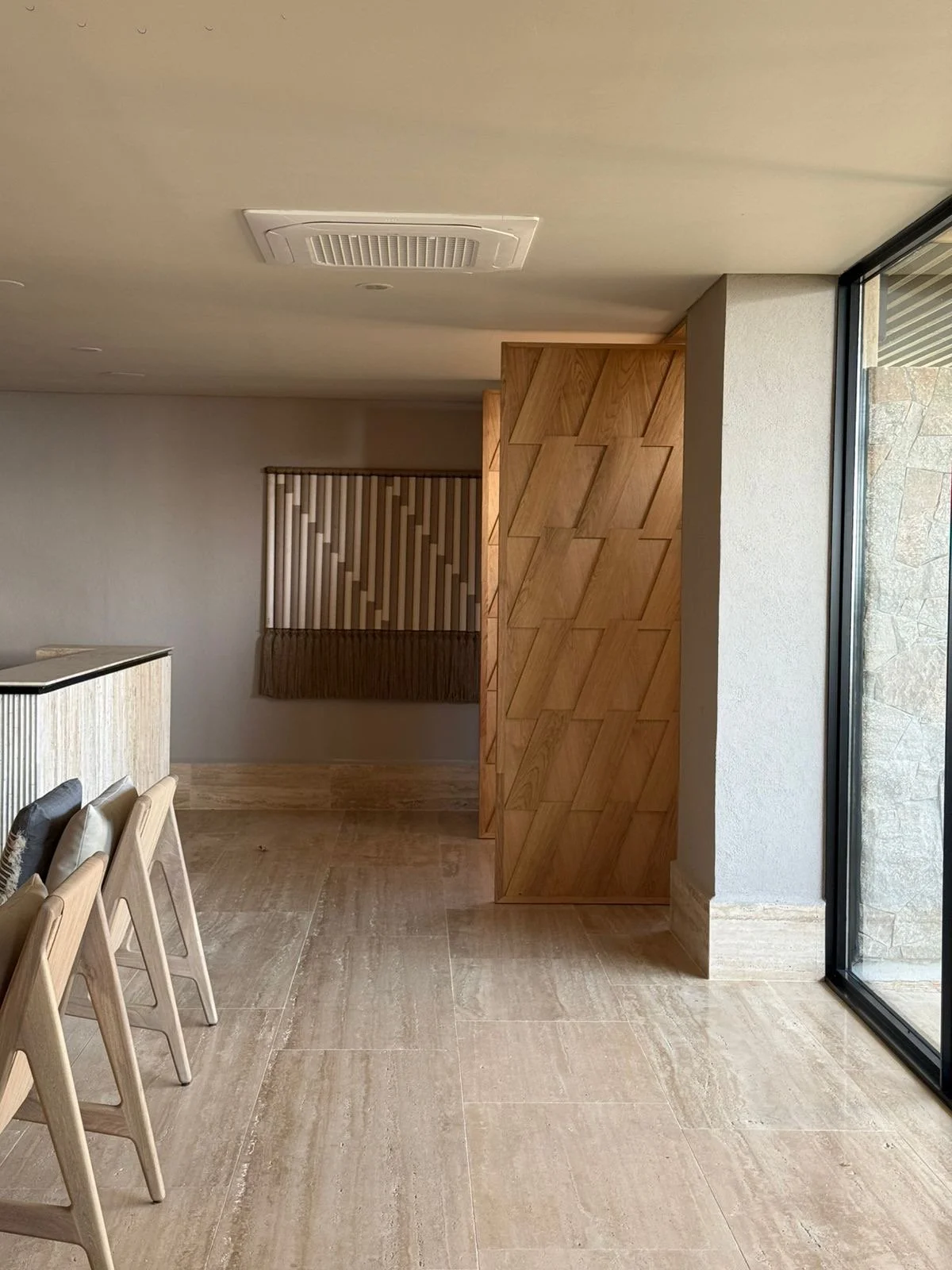 Interiores de una sala moderna con pared de piedra y madera, ventanas de vidrio, y sillones de madera con cojines.