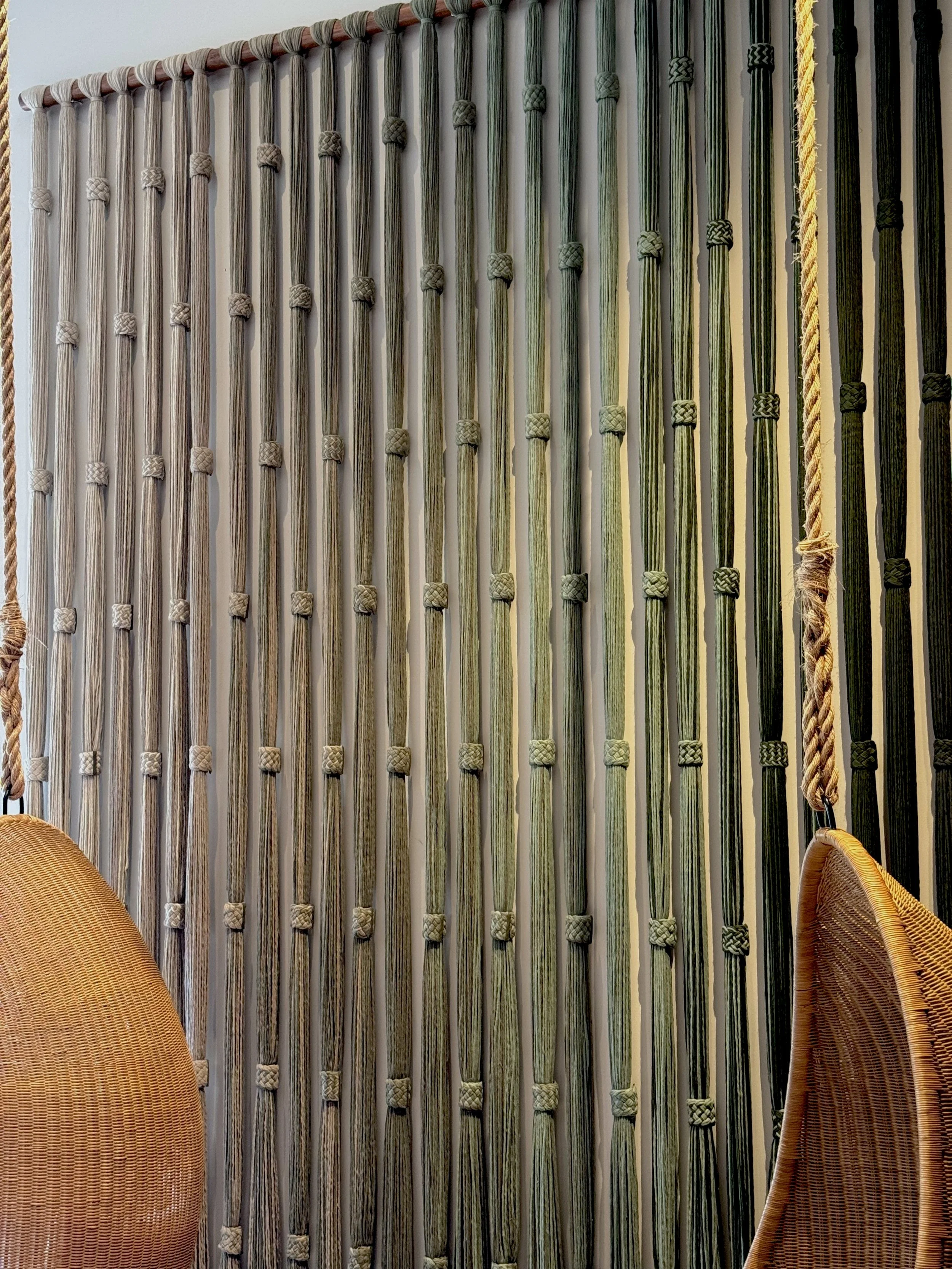 Pared decorada con cuerdas de diferentes tonos de verde y beige, entre dos sillas de mimbre en una habitación.