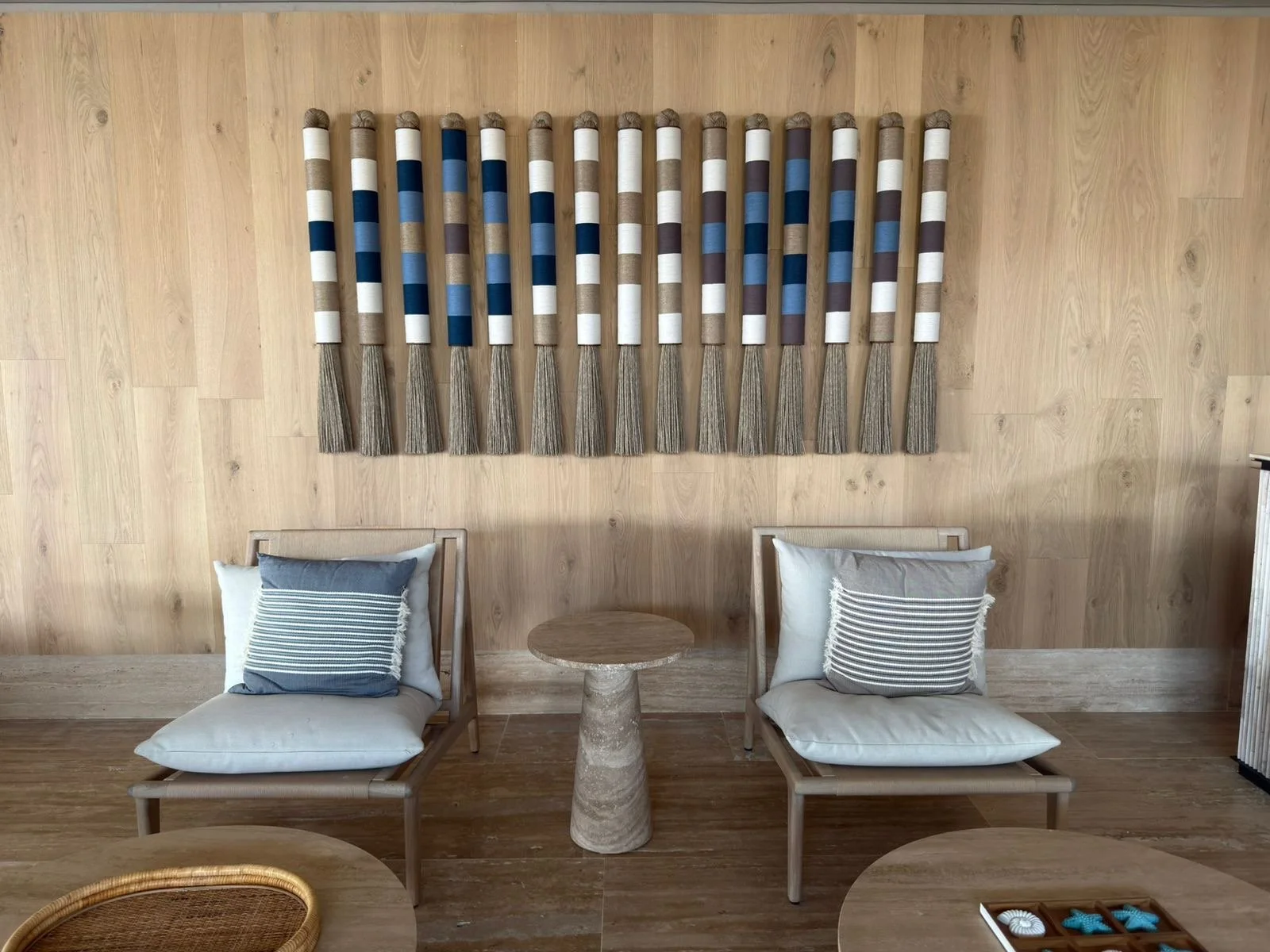 Decoración moderna con pared de madera, dos sillas de madera con cojines, una mesa entre ellas, y arte en forma de varas con bandas blancas, azules, grises y marrones en la pared.
