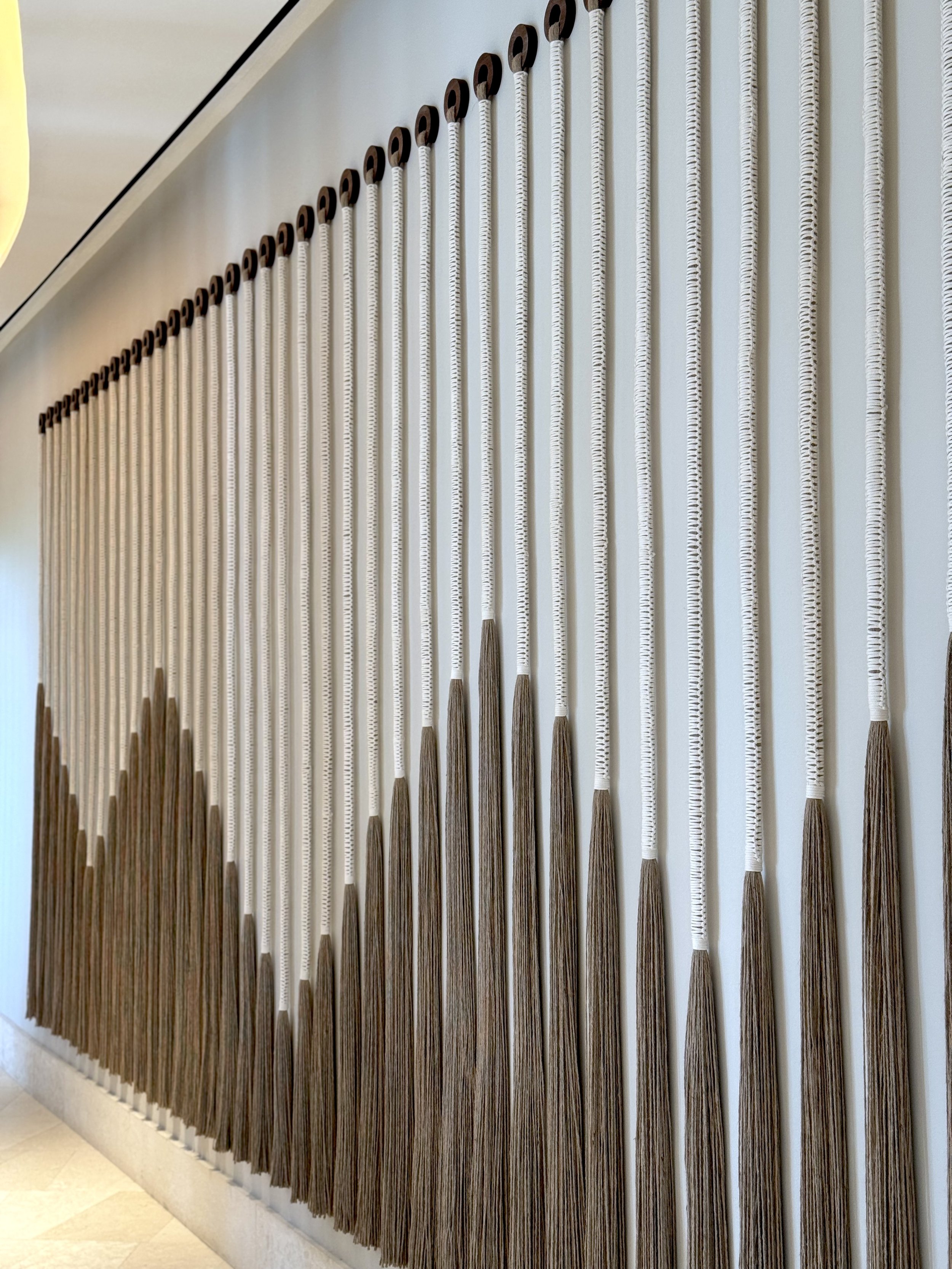 Adornos en forma de cepillos de cerdas de color blanco y cerdas de madera colgados en una pared en un patrón de líneas verticales.