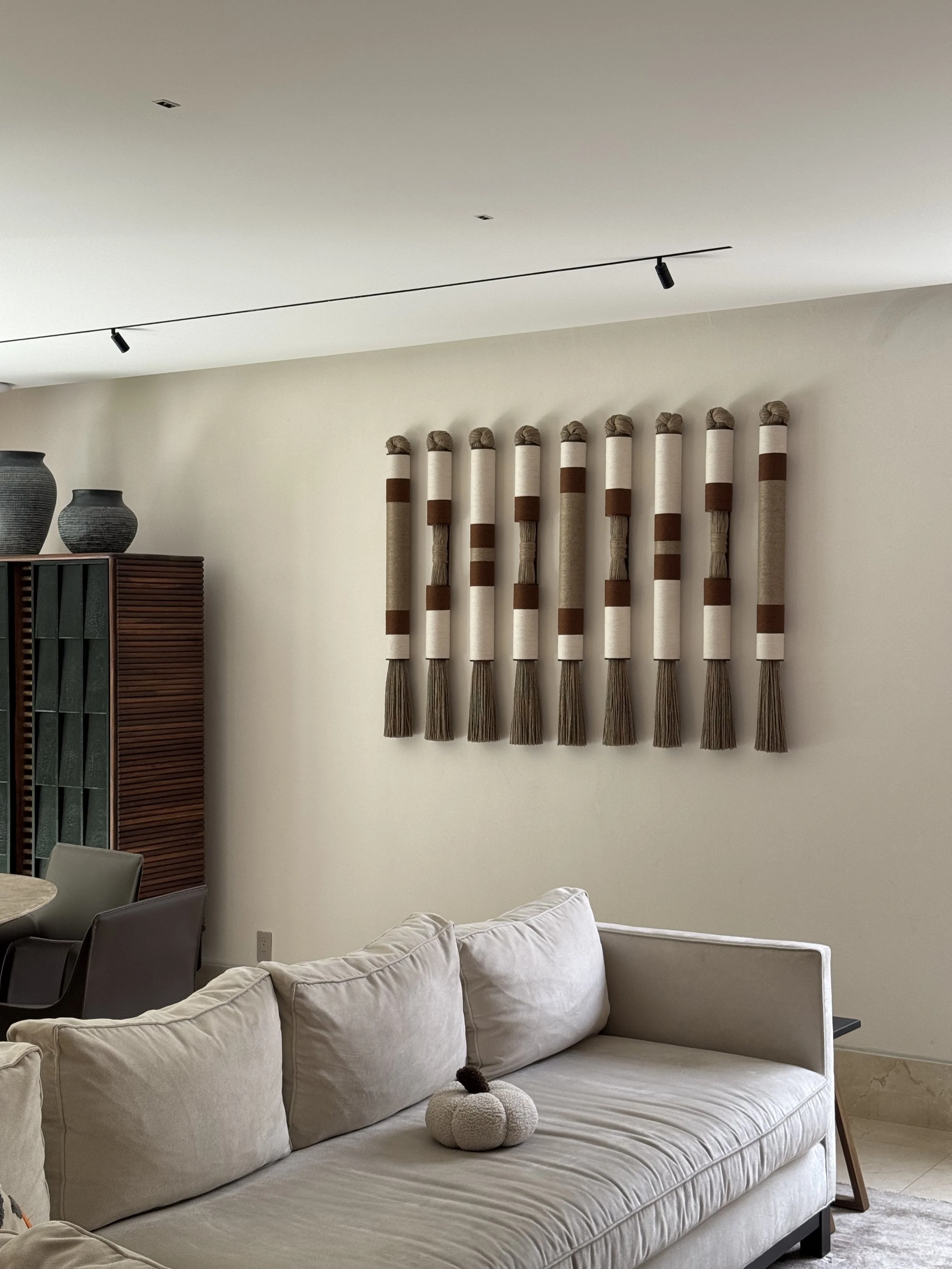 Sala con un sillón de color beige, varios cojines, una decoración en la pared con tubos decorativos de colores neutros y marrón, y dos jarrones negros sobre una estantería de madera.