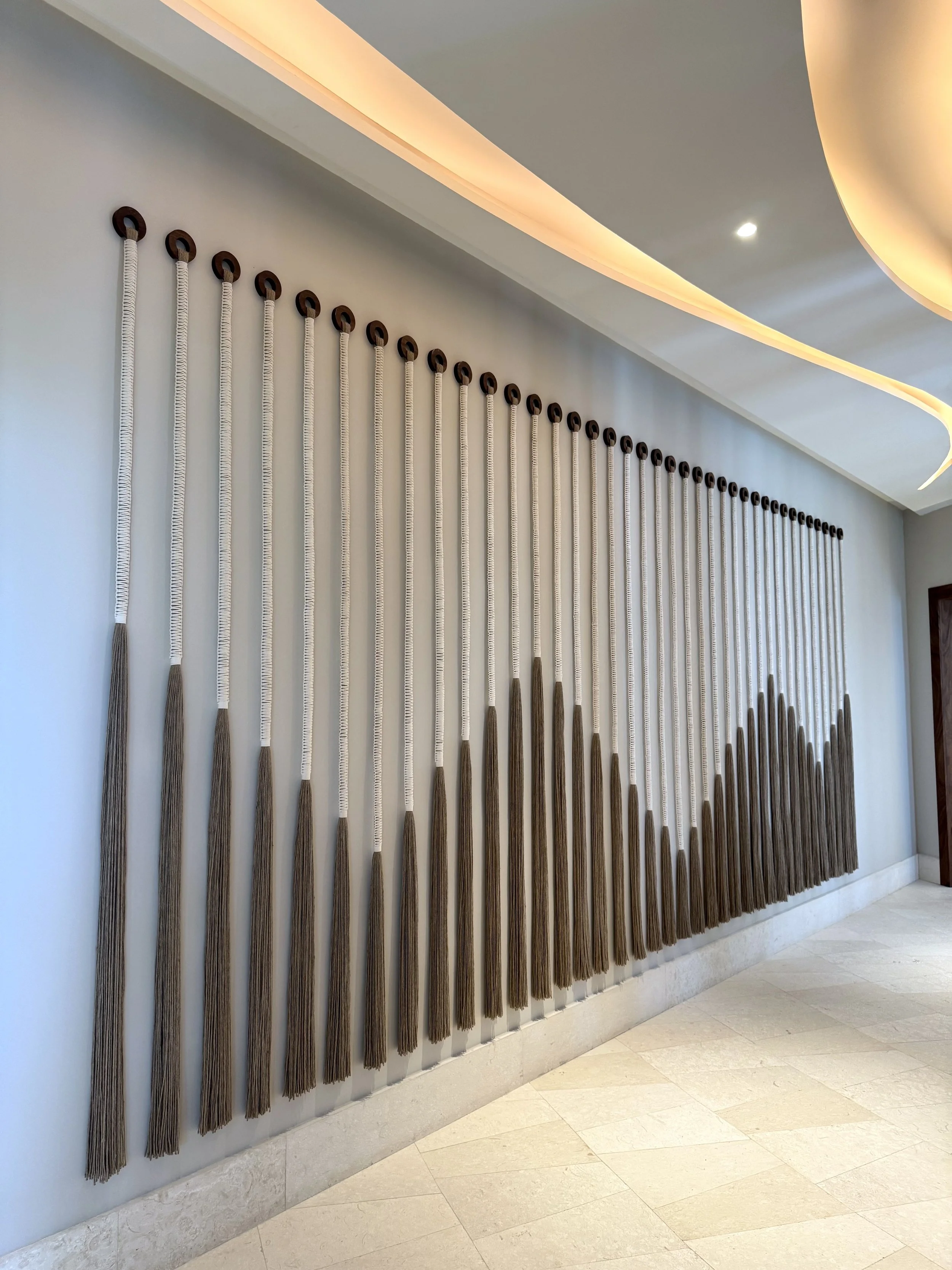 Pared decorada con hilos de colores colgados con aros de madera, en un espacio interior con techo con iluminación indirecta y piso de piedra.