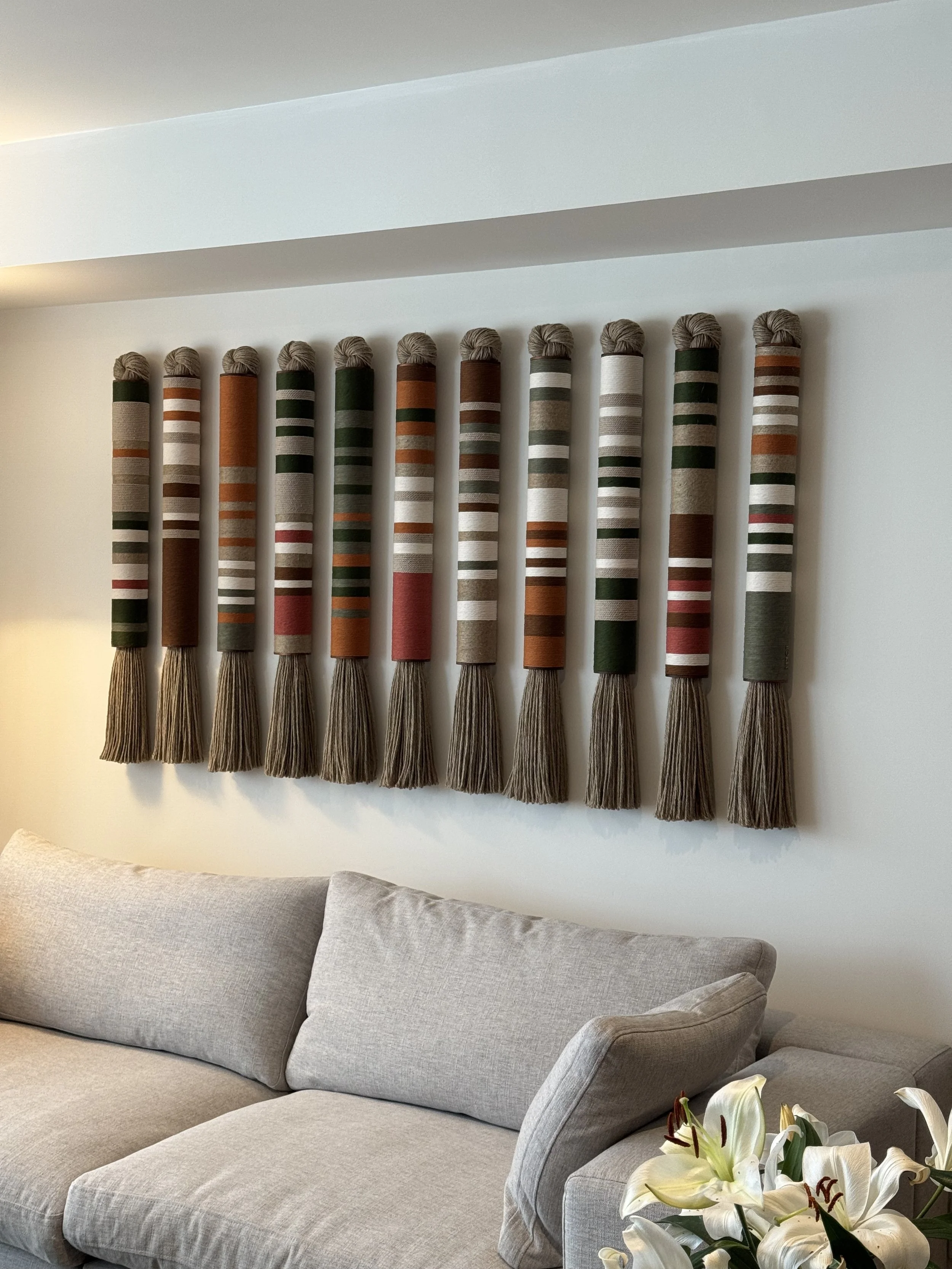 Cuatro cortinas decorativas verticales con patrones en tonos marrón, verde, gris y blanco adornan una pared, encima de un sofá de color gris en una sala moderna.