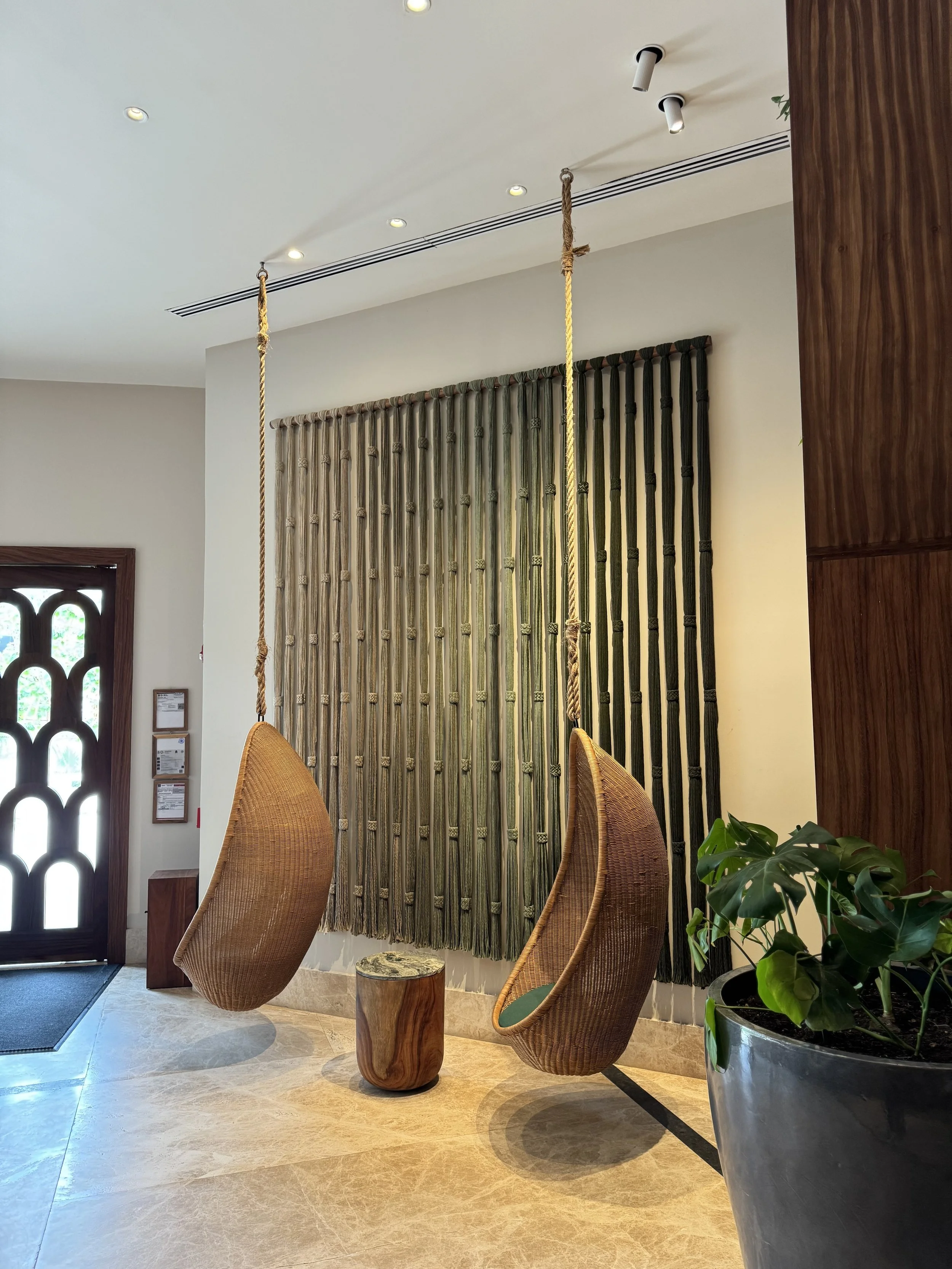 Interior de una zona de descanso con sillas colgantes de mimbre, una mesa de madera en medio y plantas decorativas, y una pared con cortinas de cuerda decorativas.