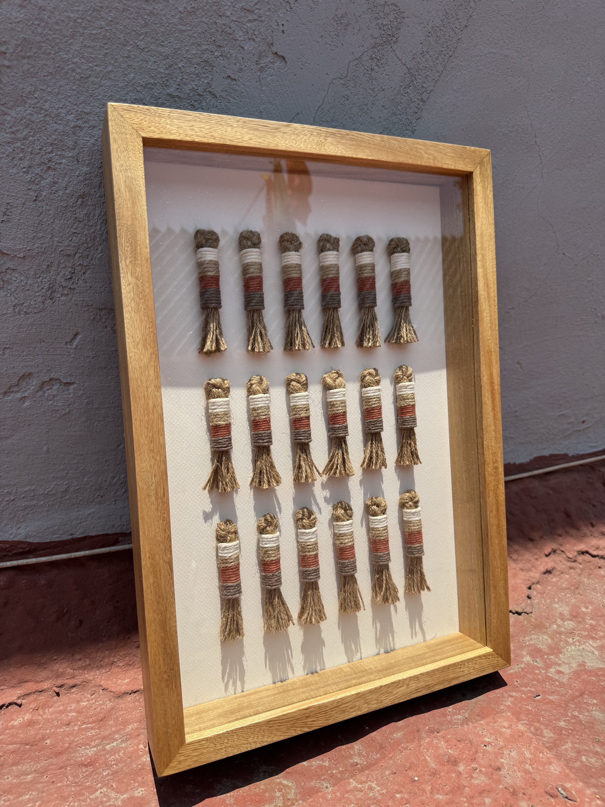 Marco de madera con hilos de colores enrollados en forma de pequeños mazos colgados en filas en una pared de color gris