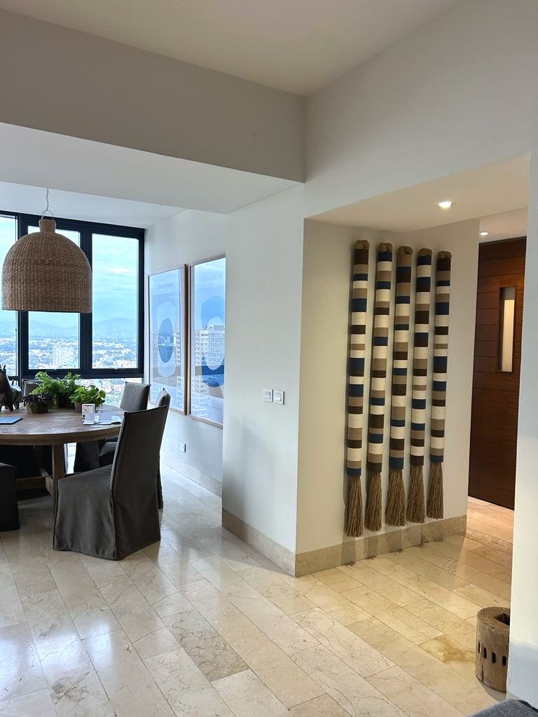 Vista de un comedor con una mesa de madera, sillas tapizadas y plantas, junto a ventanas grandes y decoraciones en las paredes, en un interior moderno y luminoso.