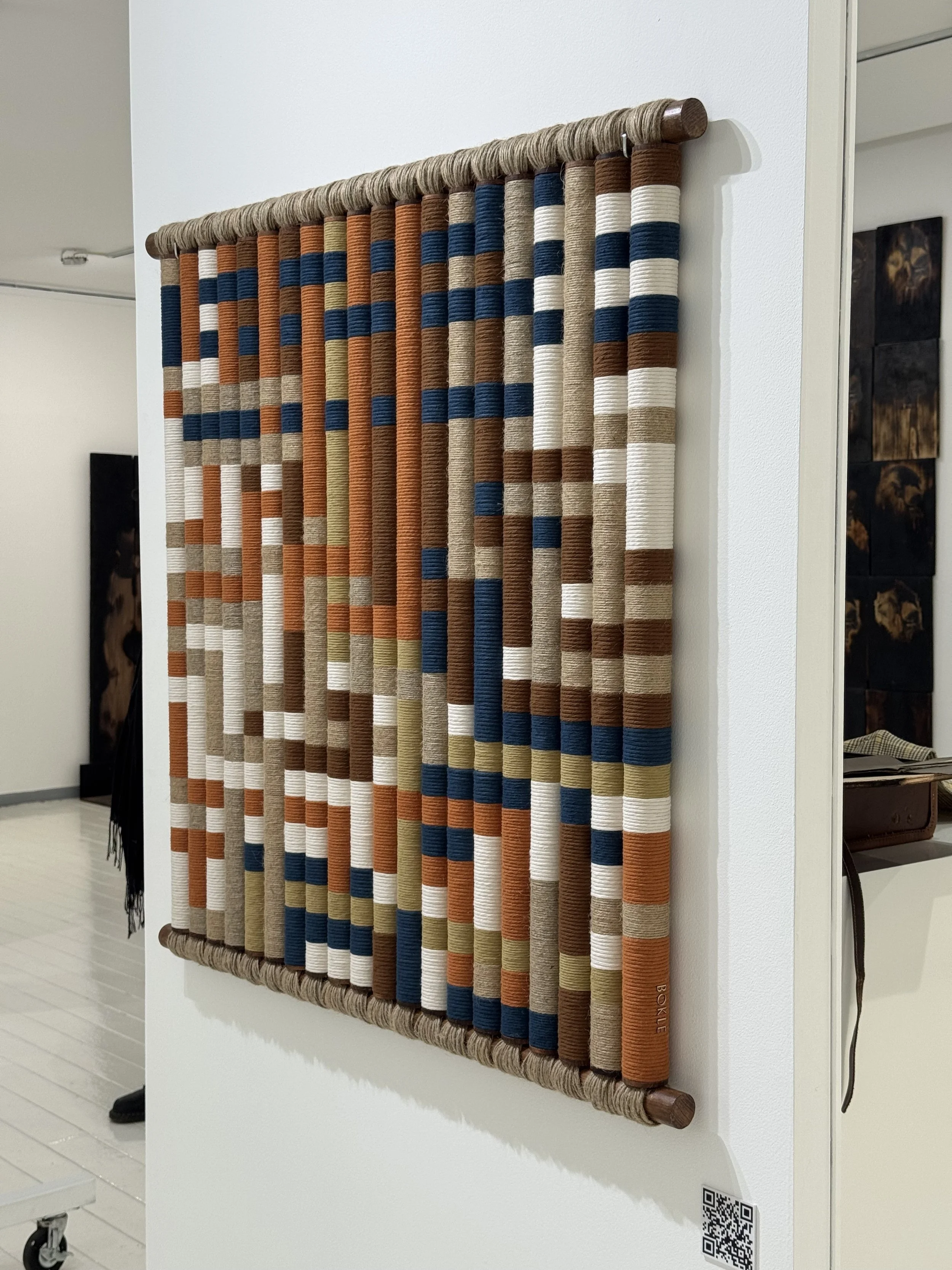 Una cortina decorativa tejida con hilos en tonos blanco, beige, azul, y marrón, instalada en una pared blanca de interior.