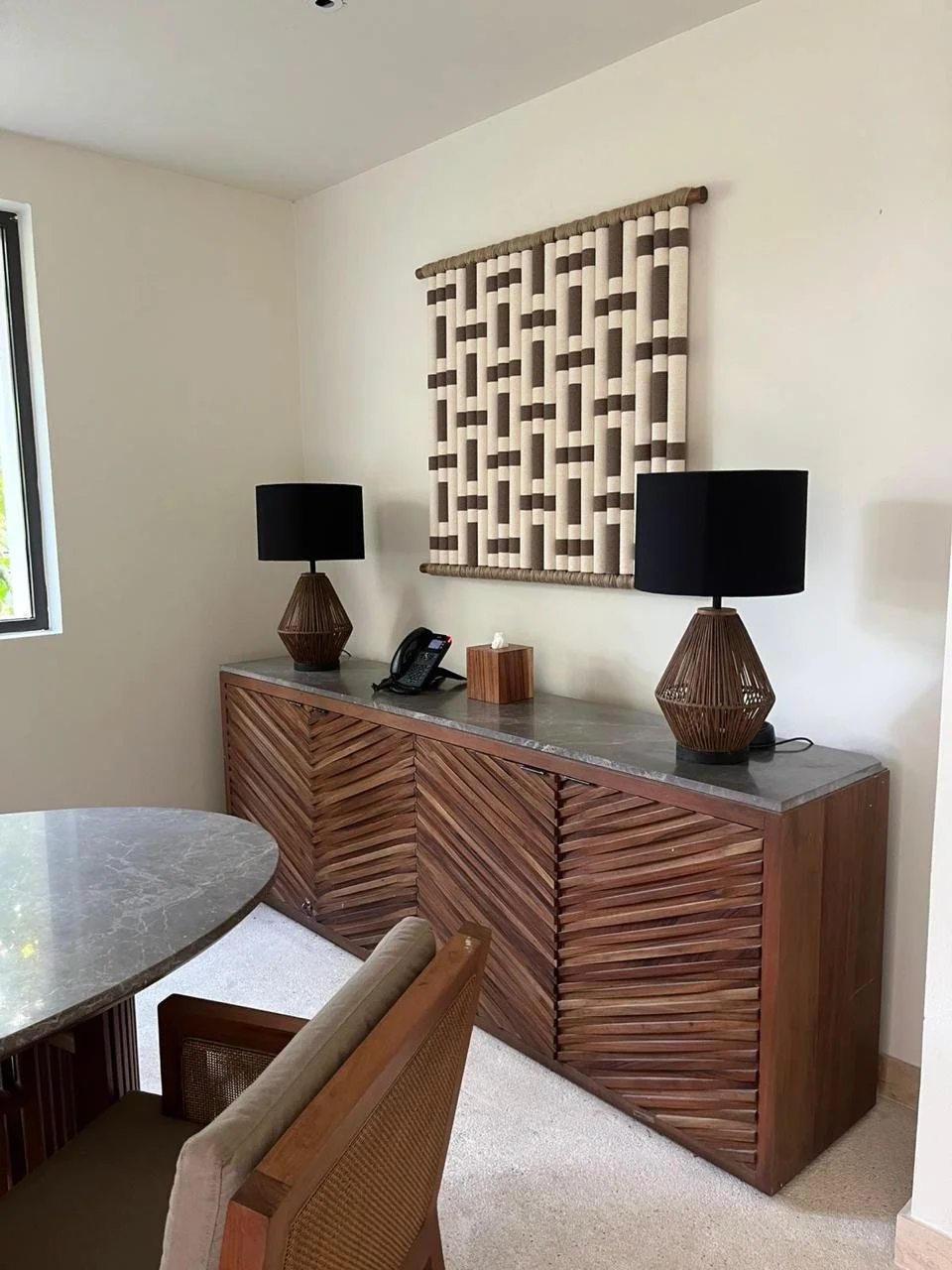Mueble de madera con puertas en zigzag y una superficie de piedra, decorado con dos lámparas negras y un teléfono en una habitación con paredes blancas y cortina beige y marrón.