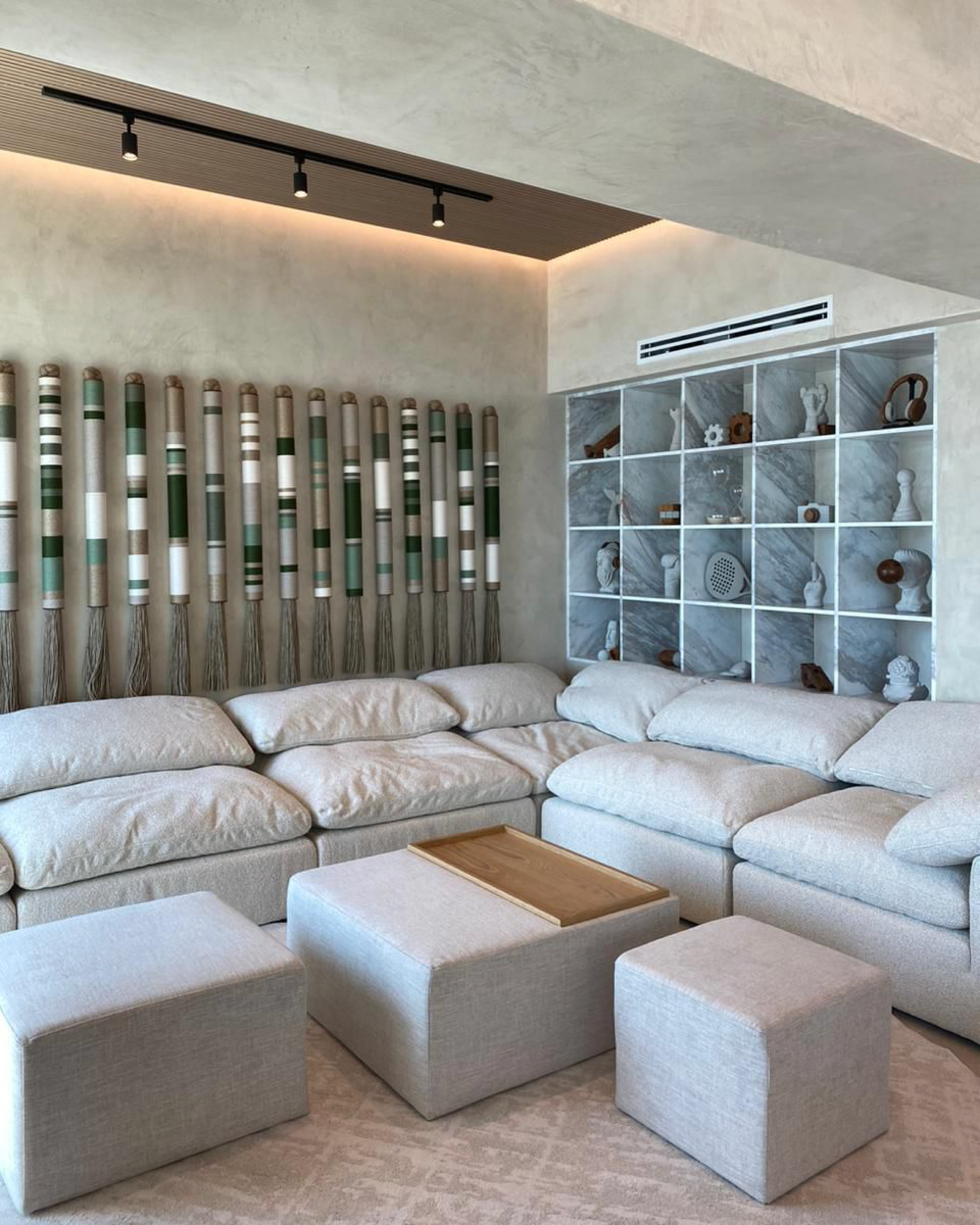 Sofá beige largo con cojines, mesas nubes pequeñas, estantería con objetos decorativos y cortinas a rayas en tonos verdes, blancos y beige, en una sala moderna.