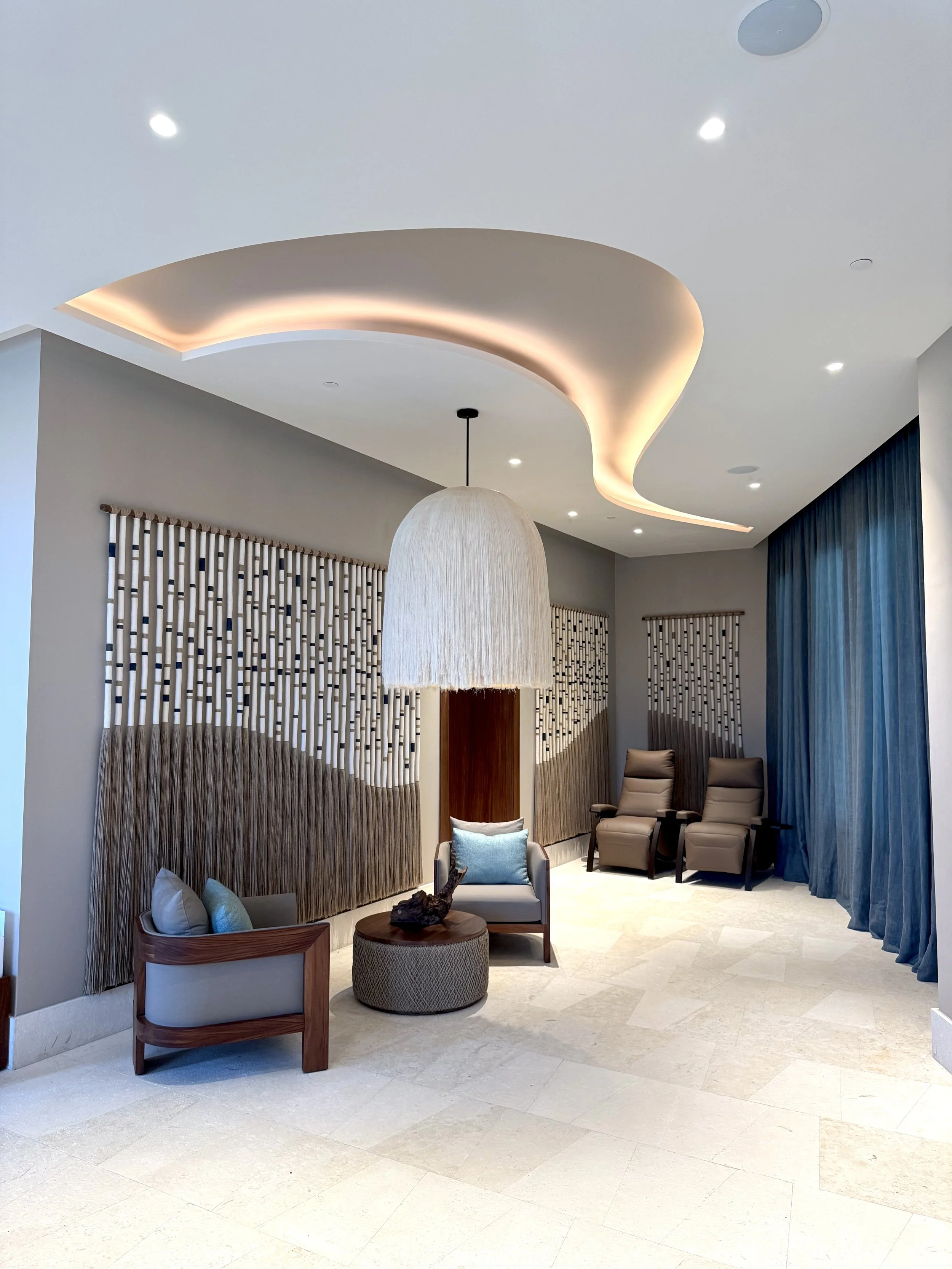 Sala de espera moderna con sillones reclinables, cortinas azules y una lámpara colgante de diseño, pisos de piedra clara y iluminación empotrada en el techo.