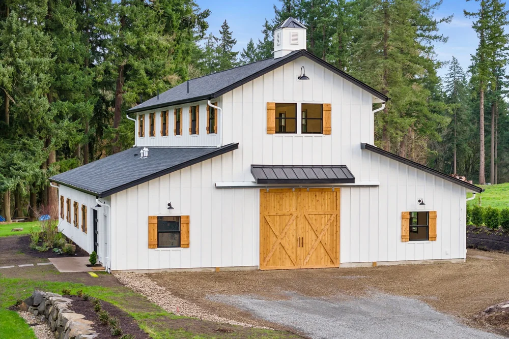 White board-and-batten barndominium with cedar shutters, large wood doors, and a roof cupola.