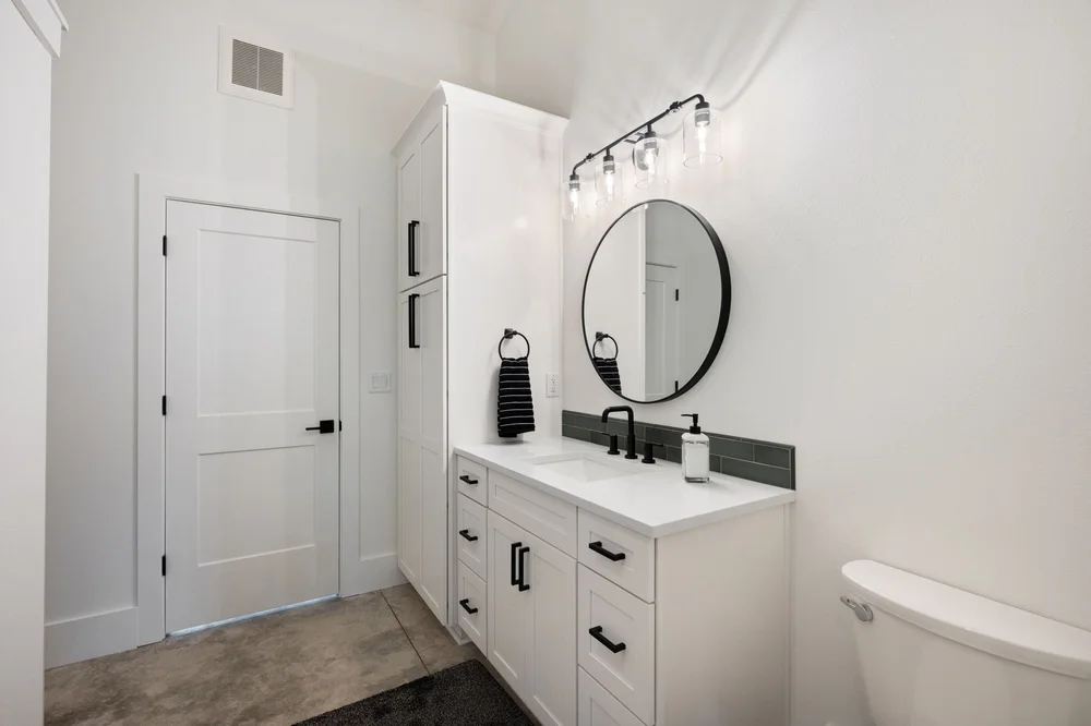Bathroom vanity with white cabinetry, a round mirror, and dark fixtures.
