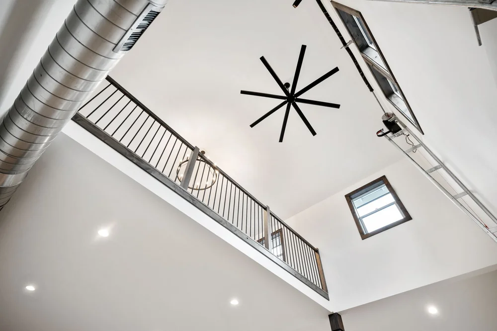 View up to the high ceiling, large fan, exposed duct, and loft railing.