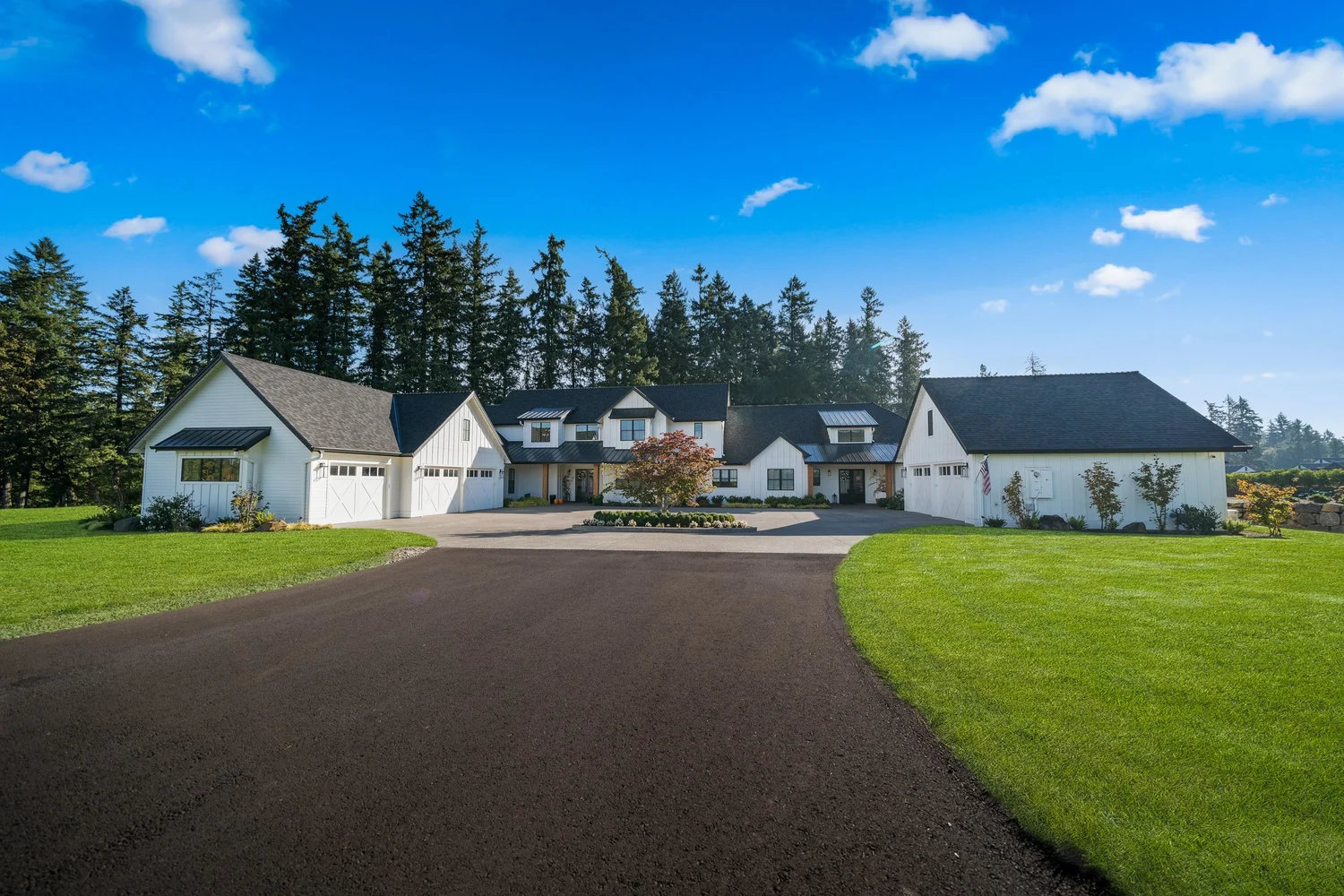 A white custom home with a circular driveway, generous lawns, and a backdrop of evergreens