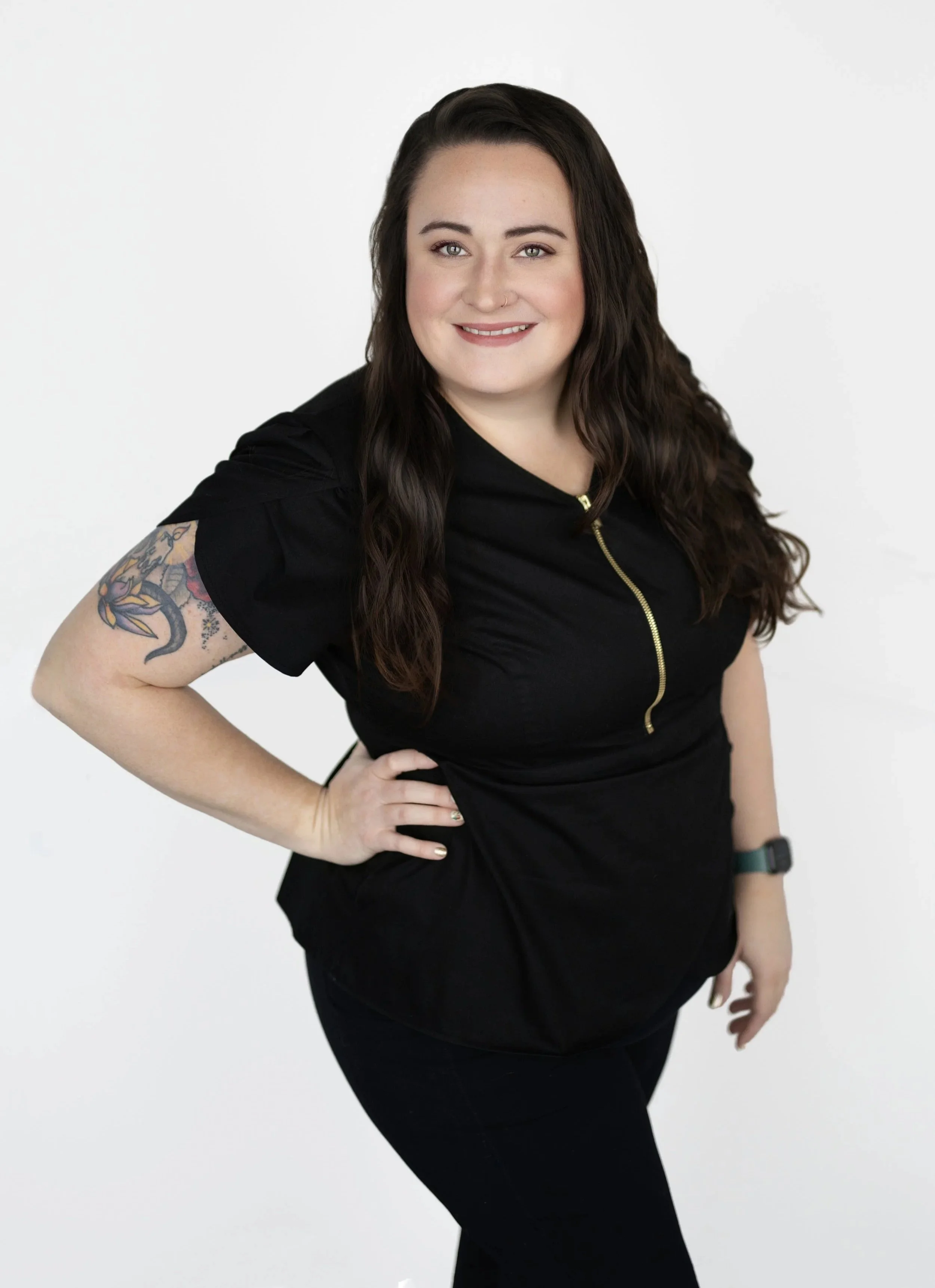 A woman with long dark wavy hair, wearing a black top with a gold zipper and black pants, standing with her hand on her hip, smiling, against a plain white background.
