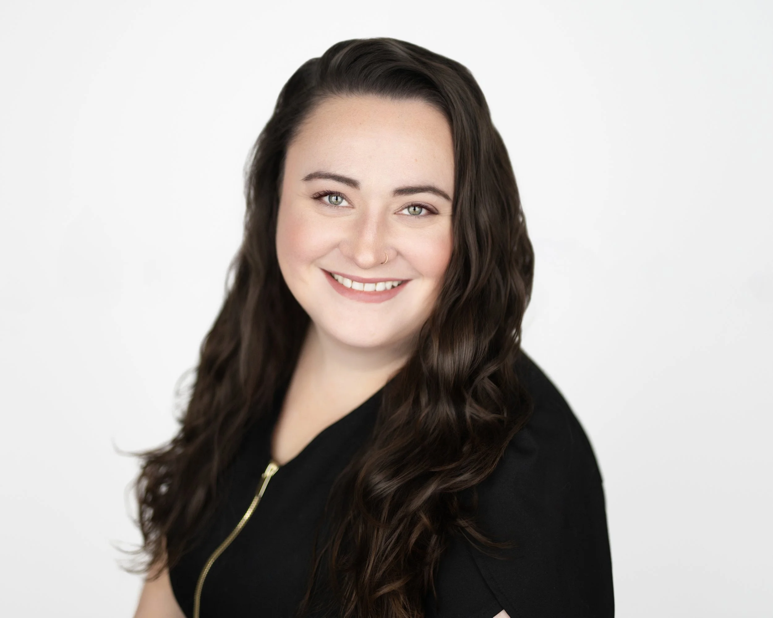 Headshot of a woman with long wavy dark hair, light skin, green eyes, smiling, wearing a black top with a gold zipper, against a plain white background.