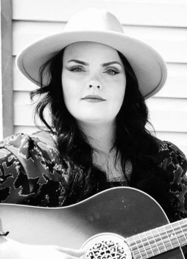 A woman with long dark hair wearing a wide brim hat and patterned blouse, holding an acoustic guitar, standing outside in front of a wooden wall.