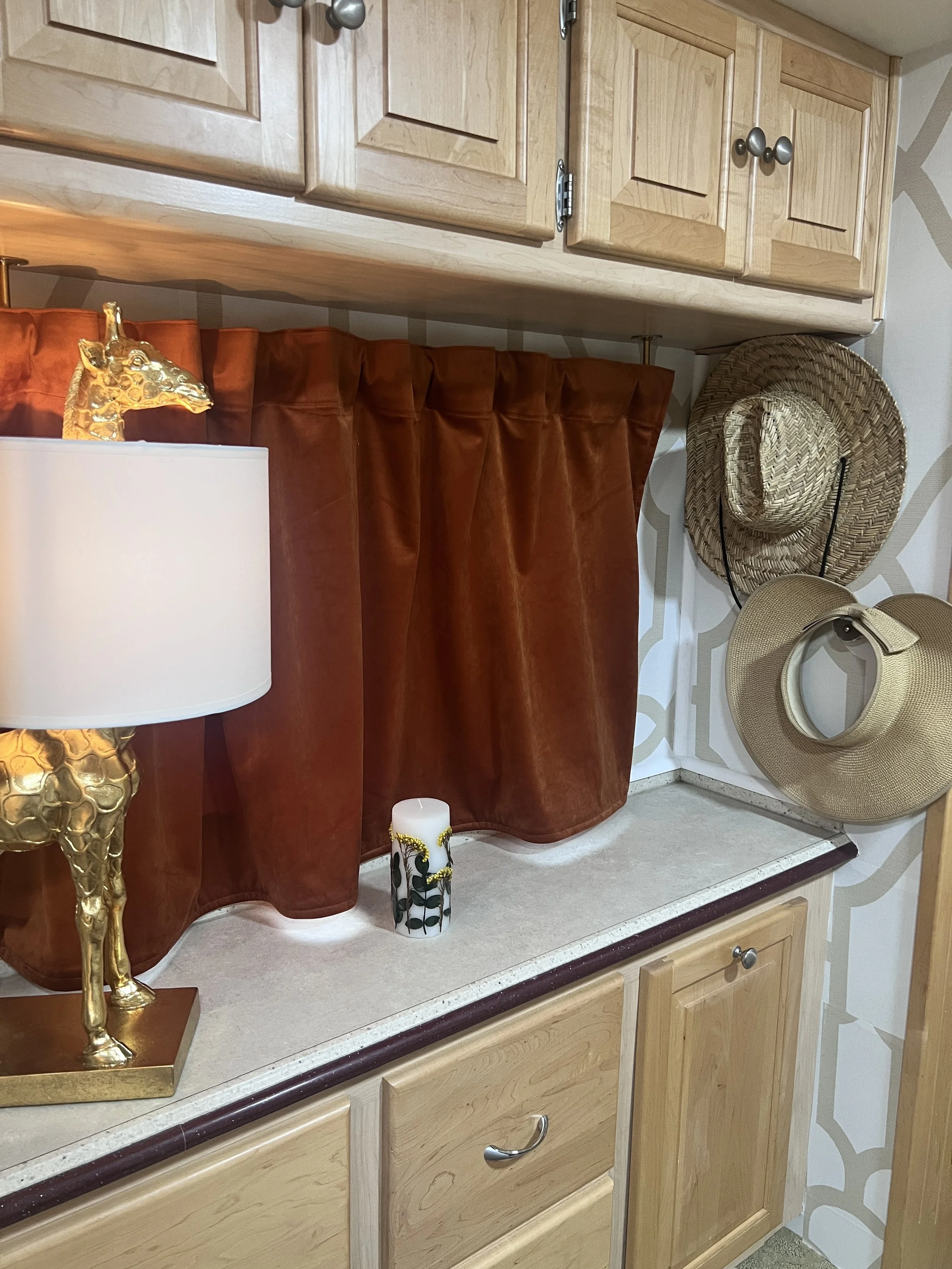 Decorative display on a kitchen or living room cabinet, featuring a lamp with a gold giraffe base, a white candle with black and gold accents, and two straw hats hanging on the wall.