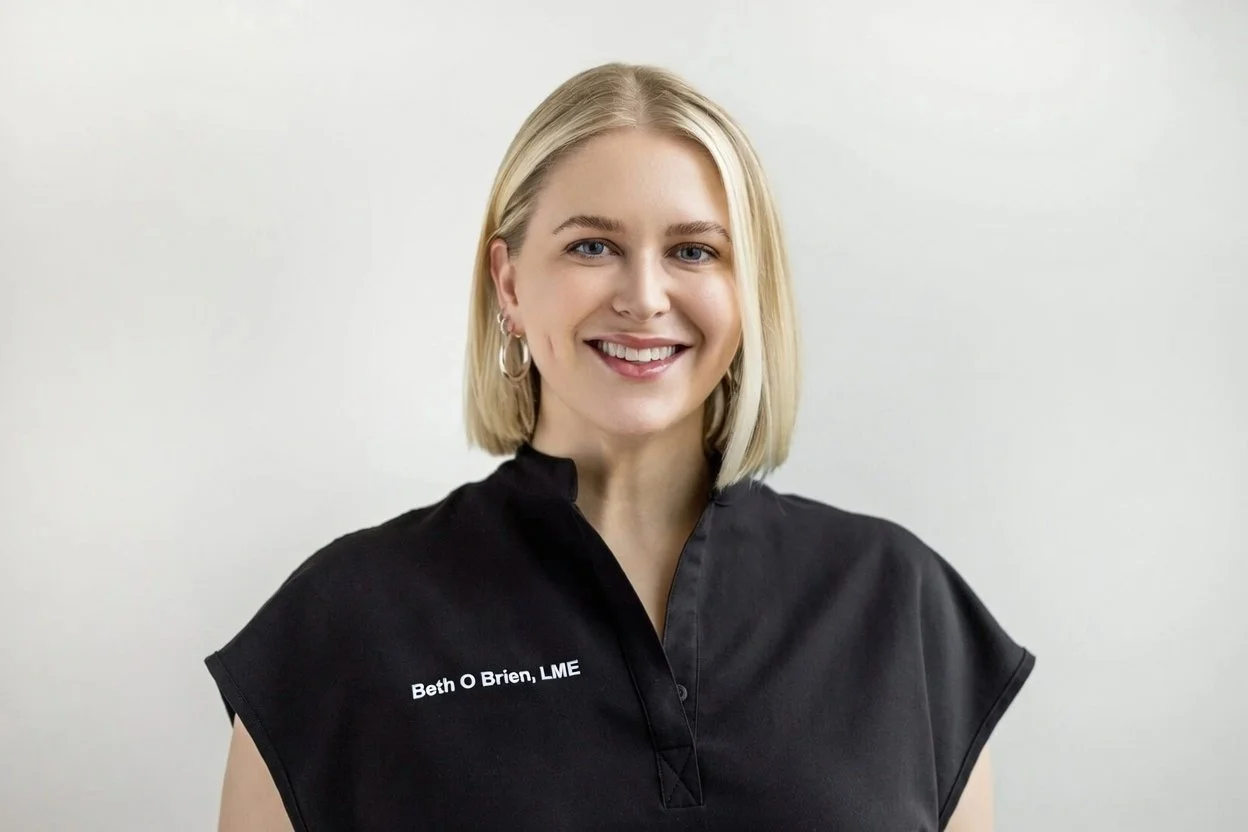 A woman with shoulder-length blonde hair and earrings smiling at the camera, wearing a black shirt with her name and credentials embroidered on it.