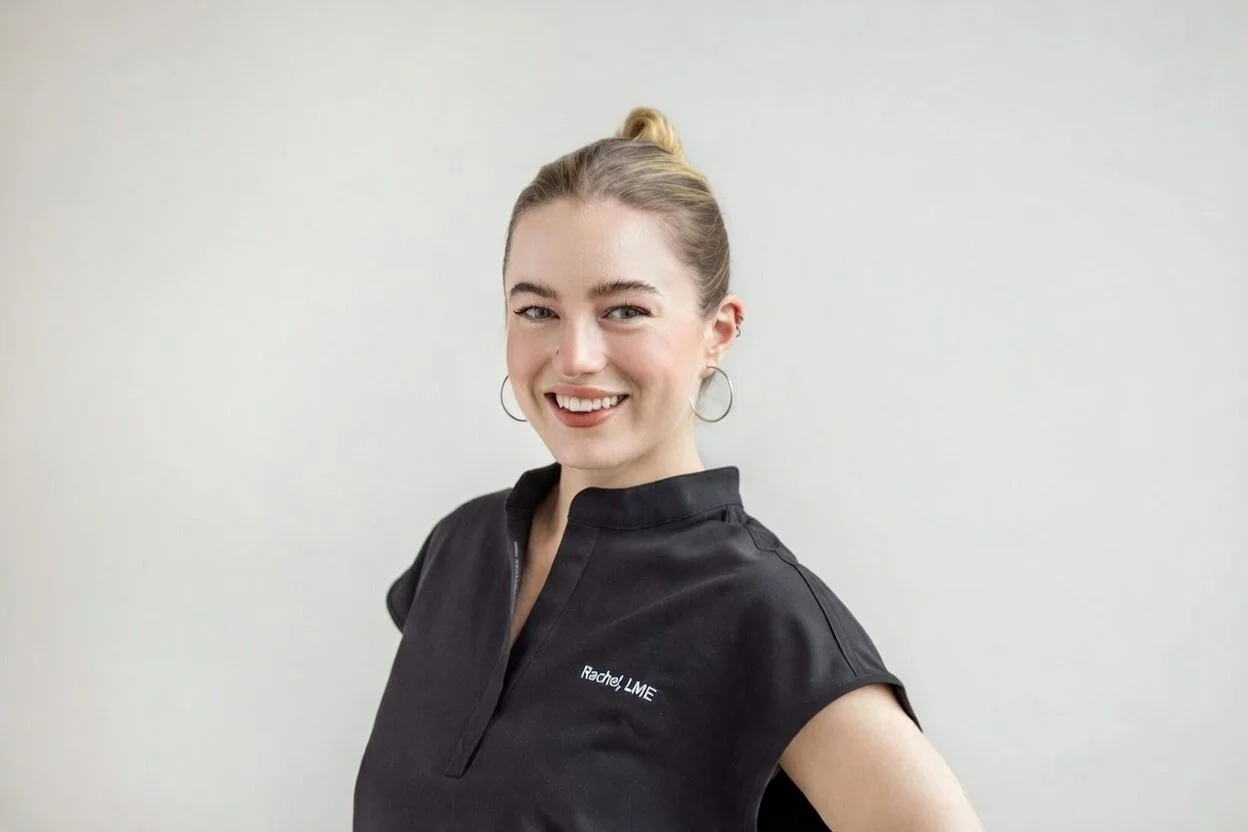 Smiling woman with blonde hair in a bun, wearing a black medical scrub top with the logo 'Rachel LME' and hoop earrings, standing against a plain light-colored wall.