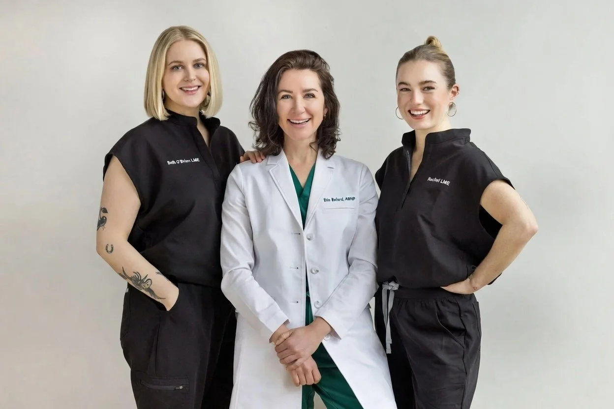 Three smiling women stand together; two in black outfits and one in a white lab coat, against a plain light background.