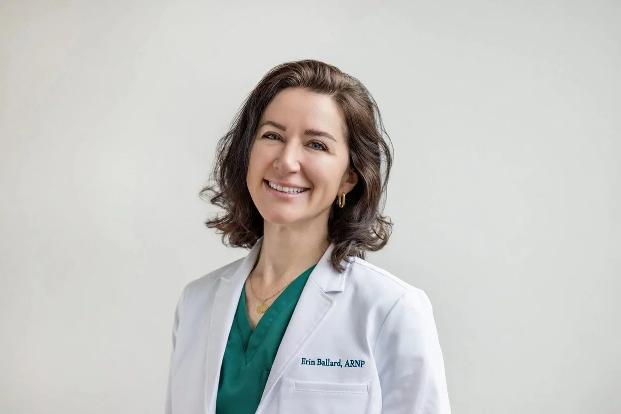 A woman in a white medical coat and green scrubs smiling against a plain background.