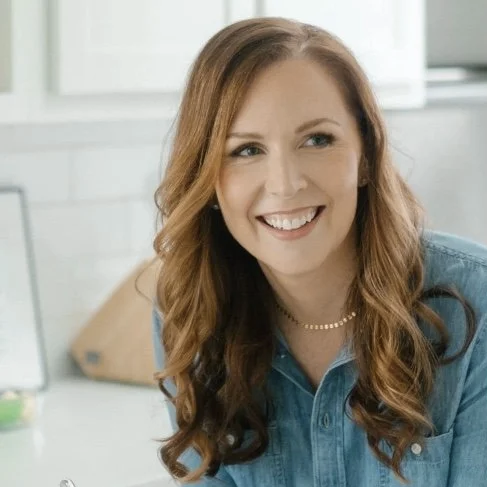A woman with long, curly red hair smiling in a kitchen setting.