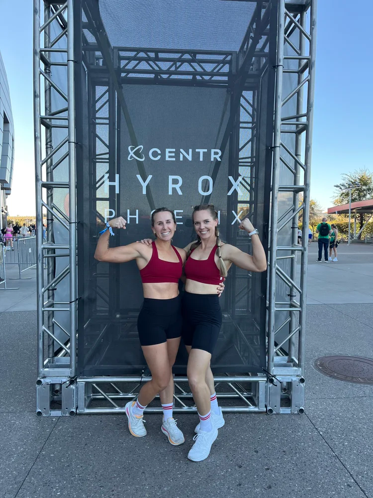 Two women in red sports bras and black shorts flexing their muscles and posing in front of a backdrop labeled 'Center Hyrox Phoenix' at an outdoor event.