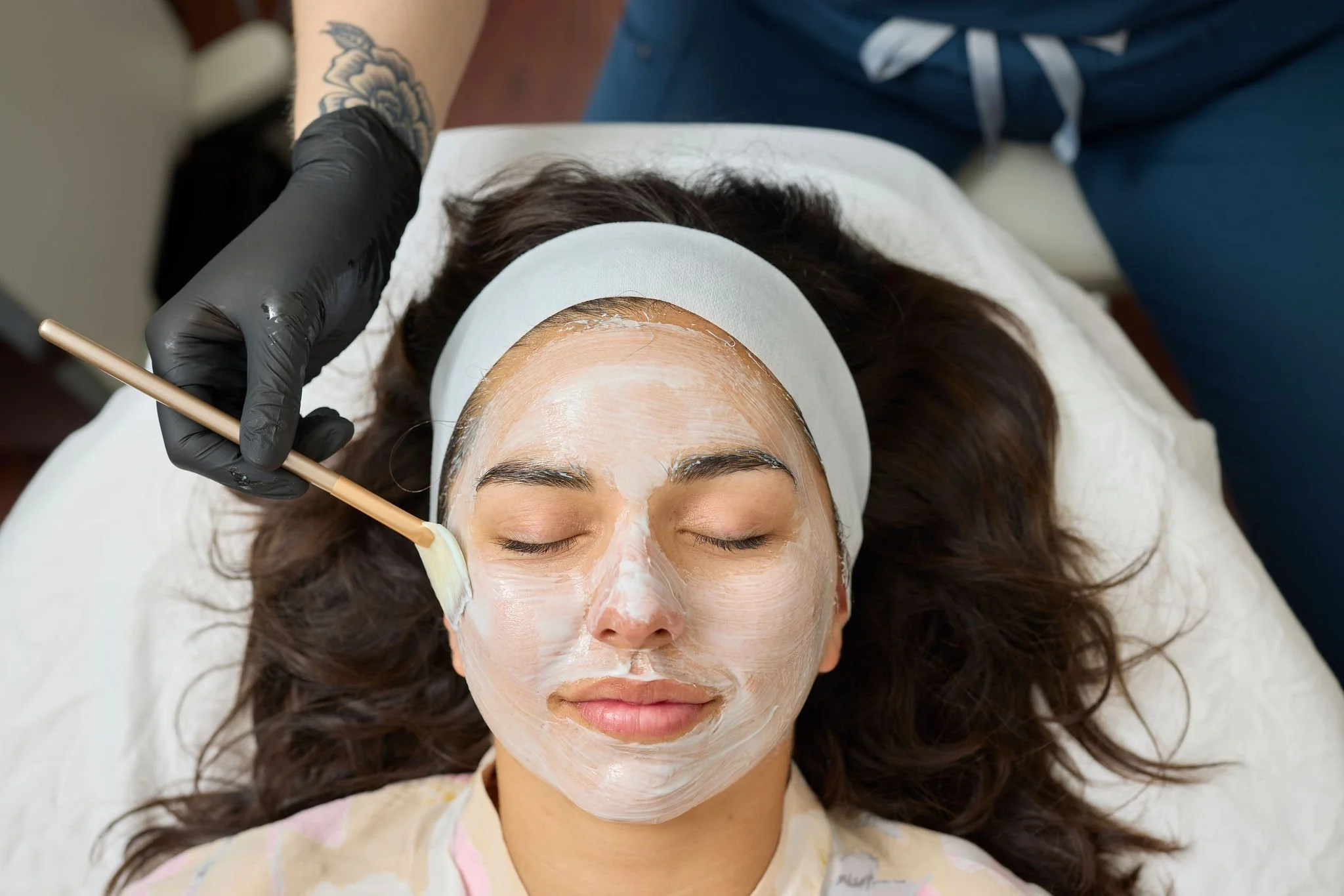 A woman with closed eyes receives a facial treatment; a brush applies a white mask to her face as she lies on a spa bed.