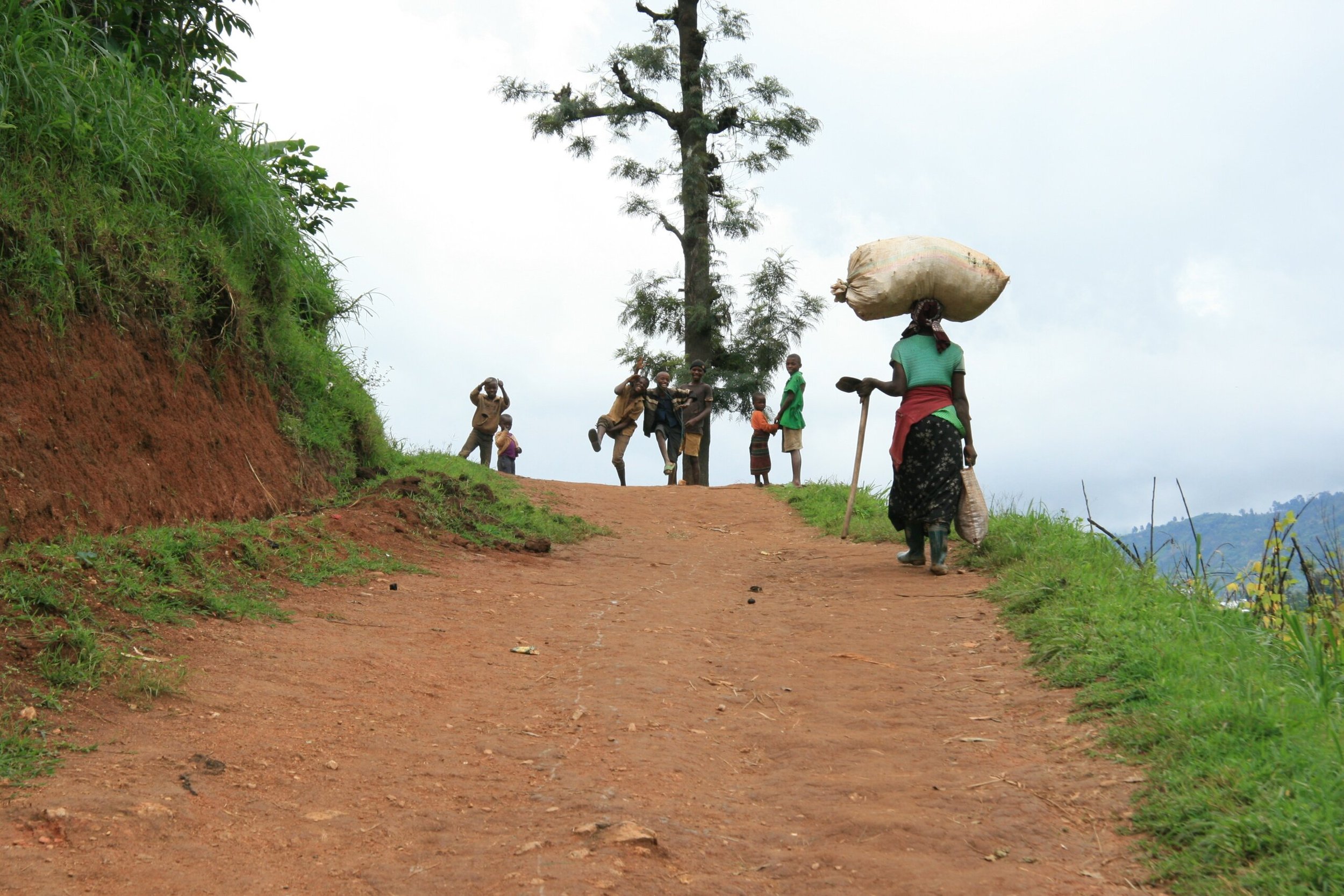 image of woman carrying sack on her head up dirt road with children playing in background