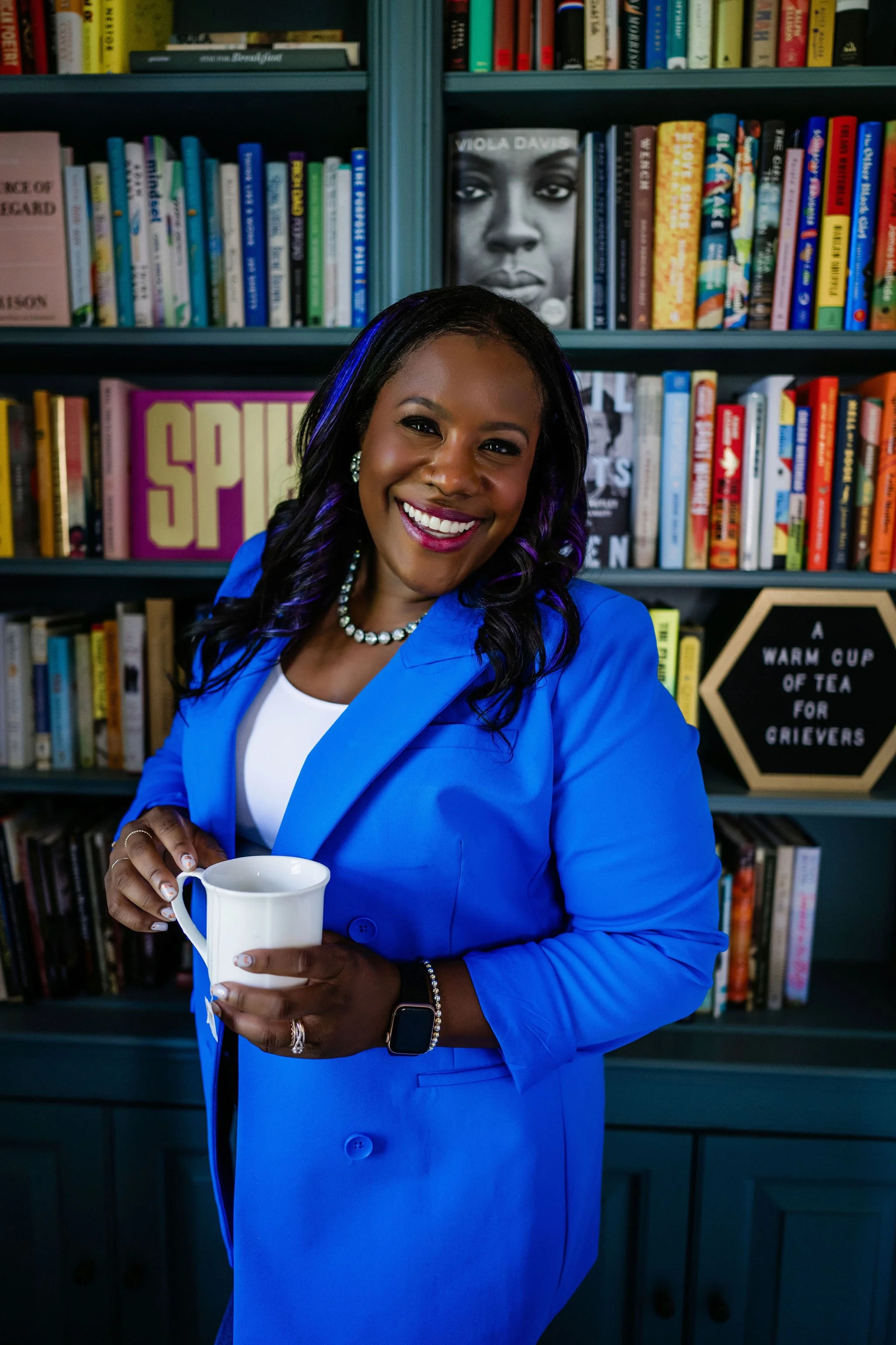 Hannah Wachter smiling while holding a mug of tea, with a "A Warm Cup of Tea for Grievers" sign visible on a bookshelf behind her