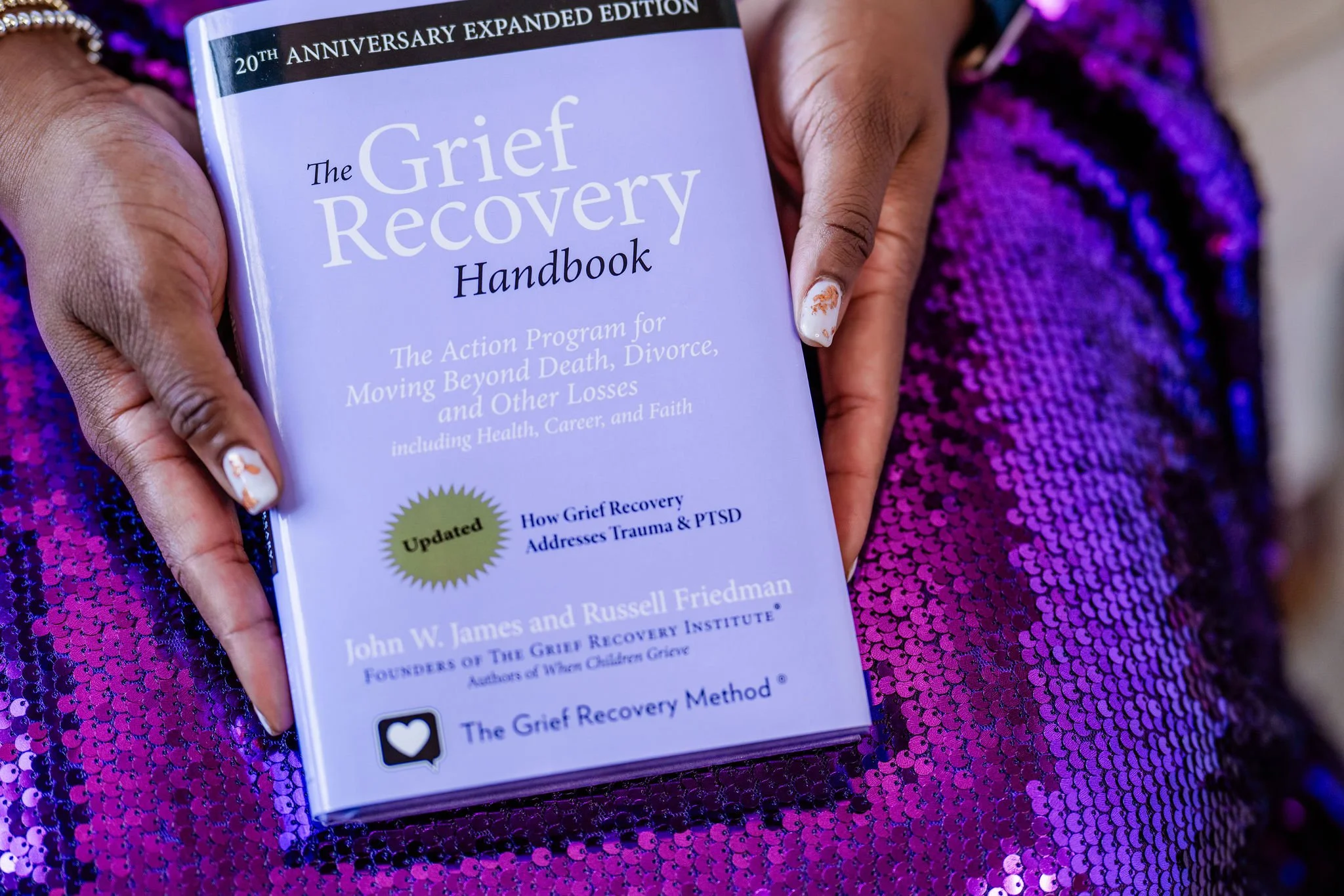 The Grief Recovery Method handbook held against a purple sequin background