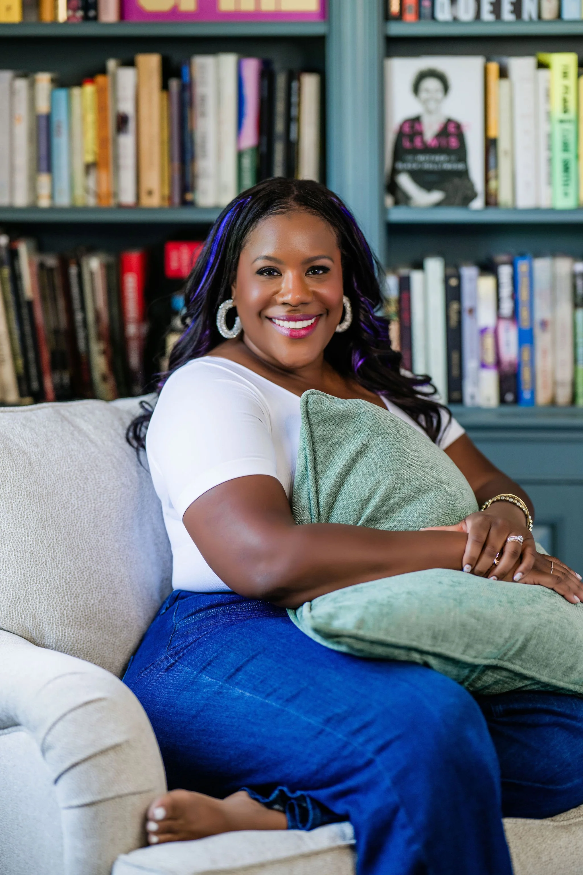 A woman smiling, sitting on a beige couch with a green pillow, in a room with bookshelves filled with books in the background.