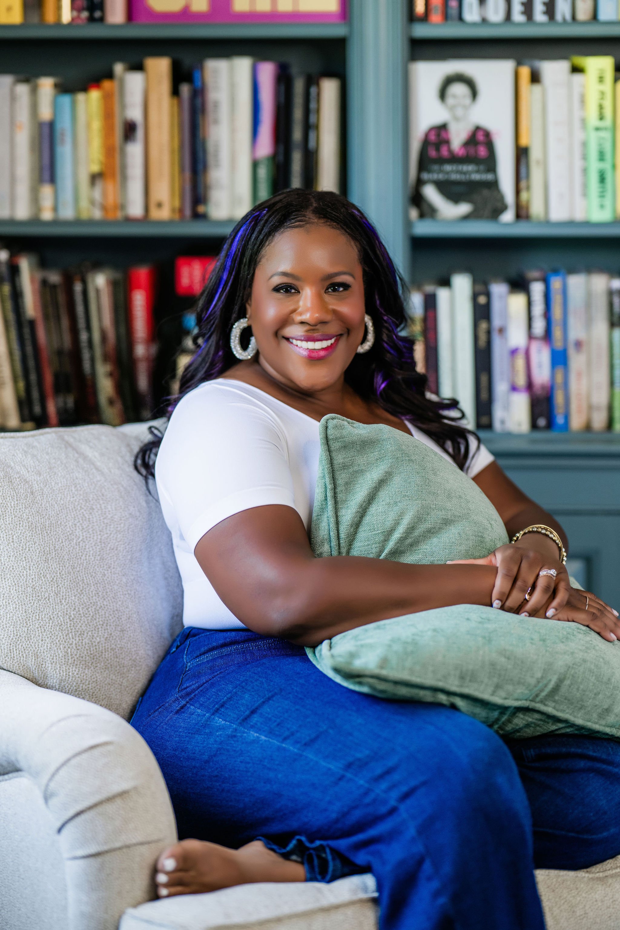 A woman smiling, sitting on a beige couch with a green pillow, in a room with bookshelves filled with books in the background.
